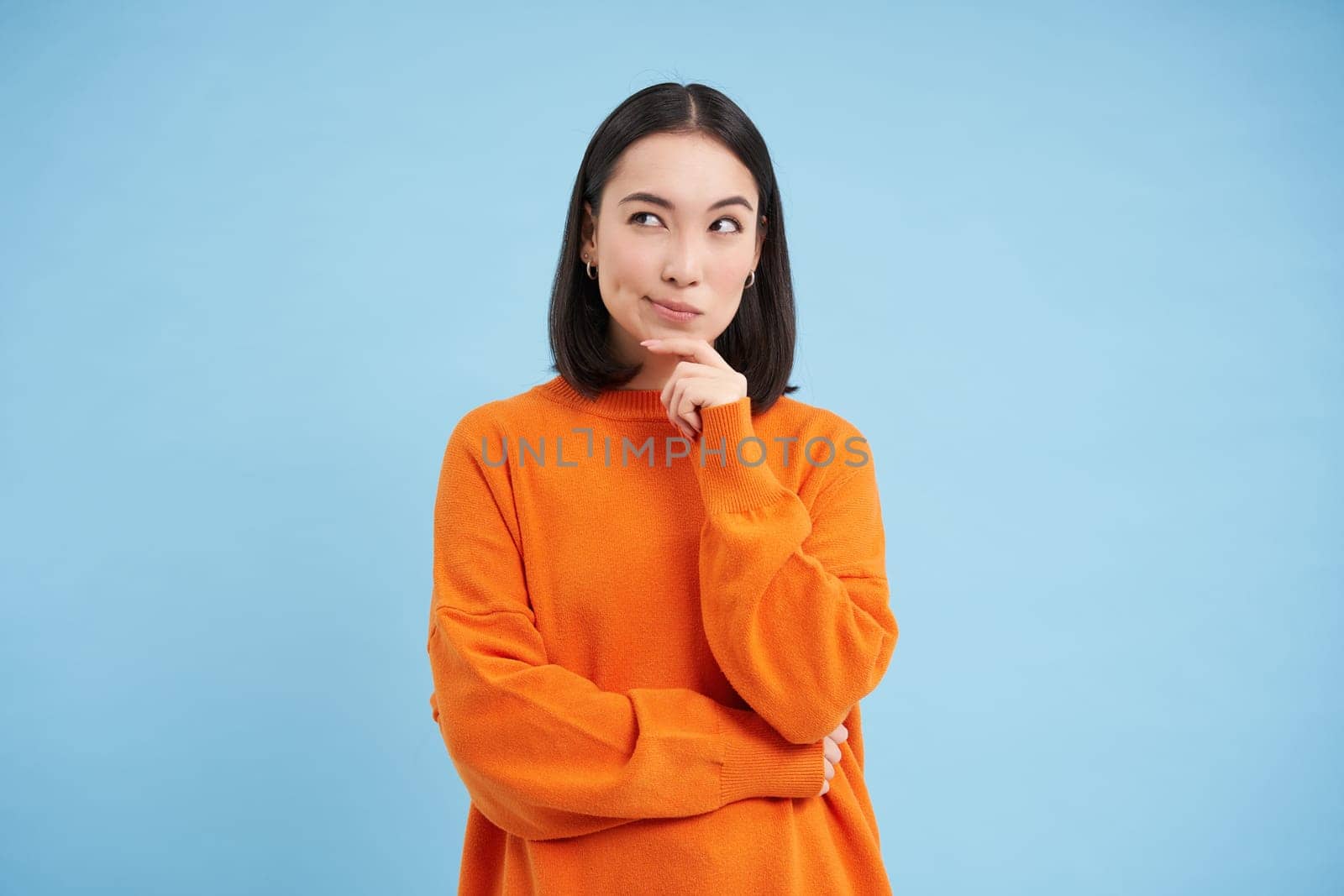 Brainstorm, ideas concept. Portrait of asian girl thinks and looks puzzled, stands thoughtful in orange sweatshirt, makes decision, blue background.