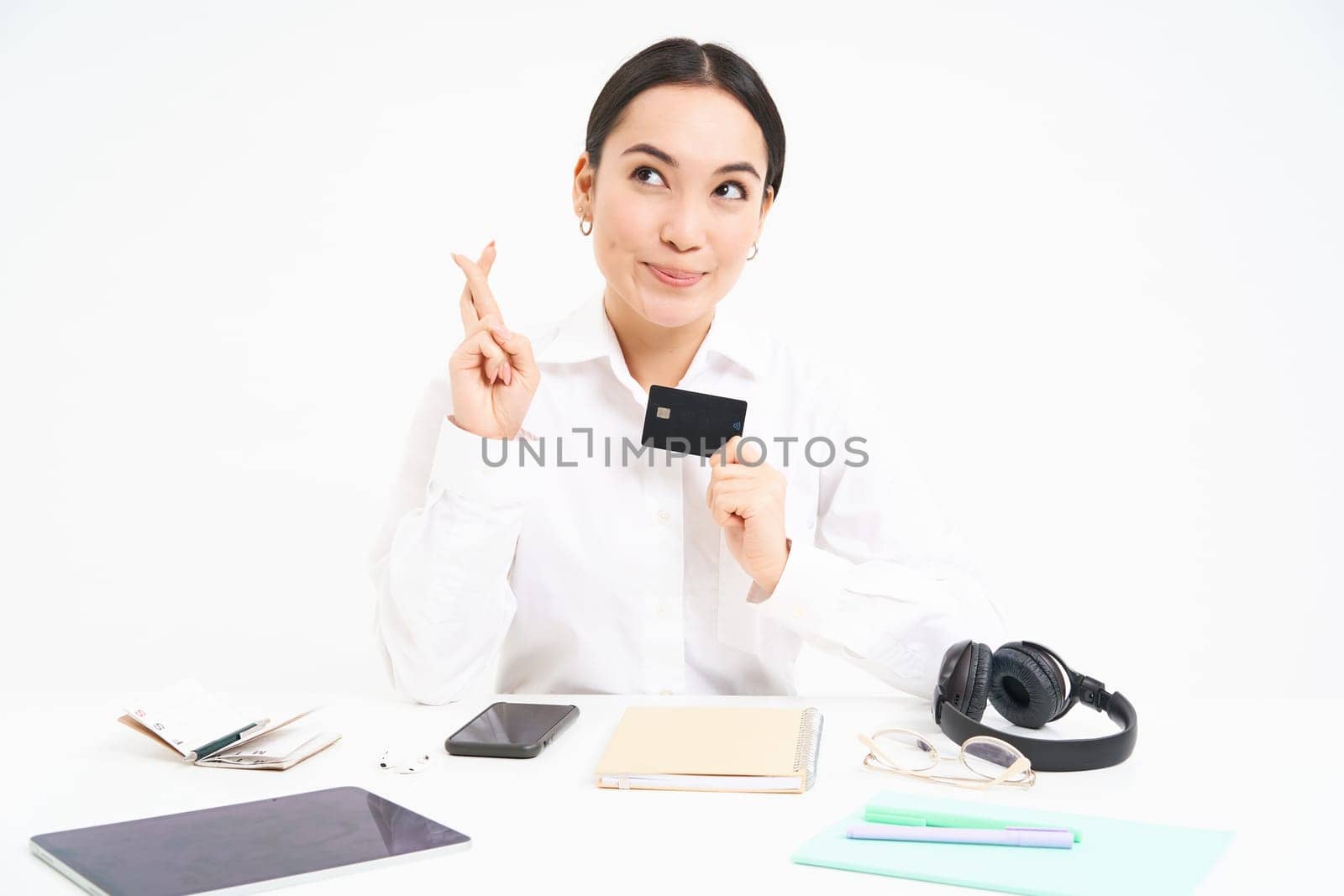 Hopeful asian woman, office worker shows credit card and cross fingers for good luck, makes wish, white background.