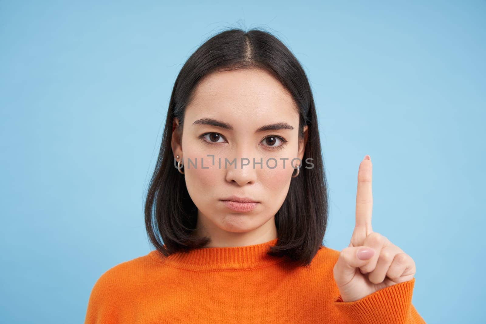 Close up of serious asian woman shows one finger, stop gesture, scolding, disapprove smth, stands over blue background by Benzoix