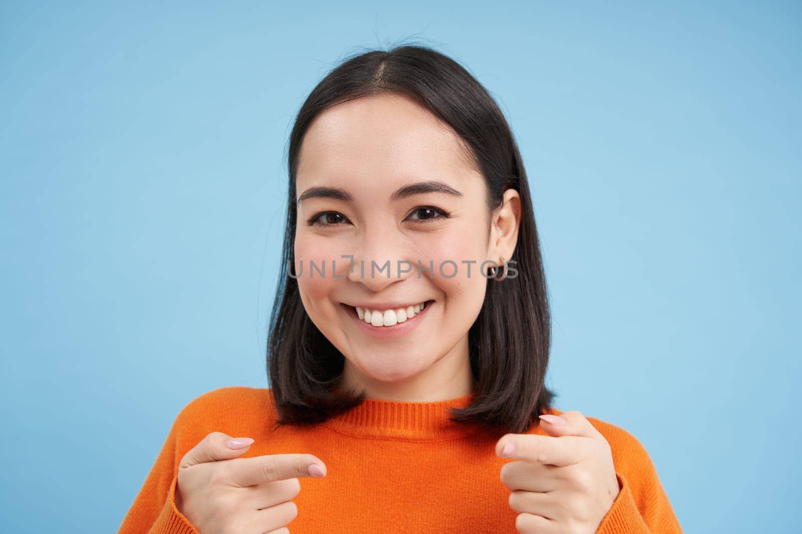 Gotcha. Close up portrait of smiling asian woman points fingers at camera pleased, congratulates you, stands over blue background by Benzoix