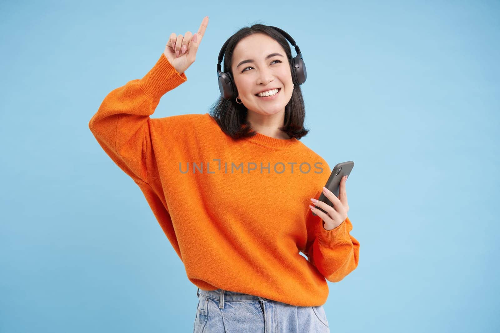 Beautiful korean woman in headphones, dancing, listening music on mobile phone app, smiling, posing over blue background.