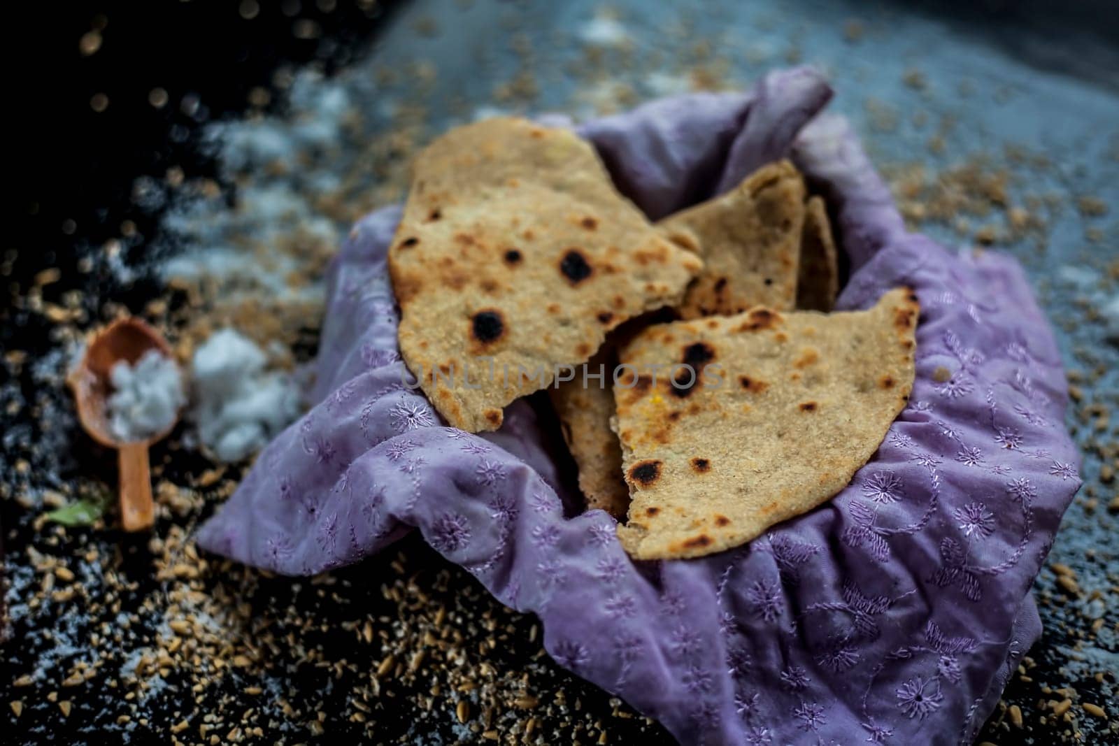 Close-up shot of round bread Bhakri on the black wooden surface along with some raw whole wheat, and salt in a container. Shot of Bhakri in a container on the black surface.