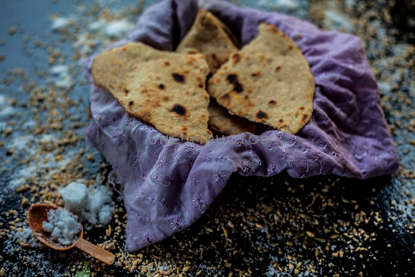Close-up shot of round bread Bhakri on the black wooden surface along with some raw whole wheat, and salt in a container. Shot of Bhakri in a container on the black surface. by mirzamlk