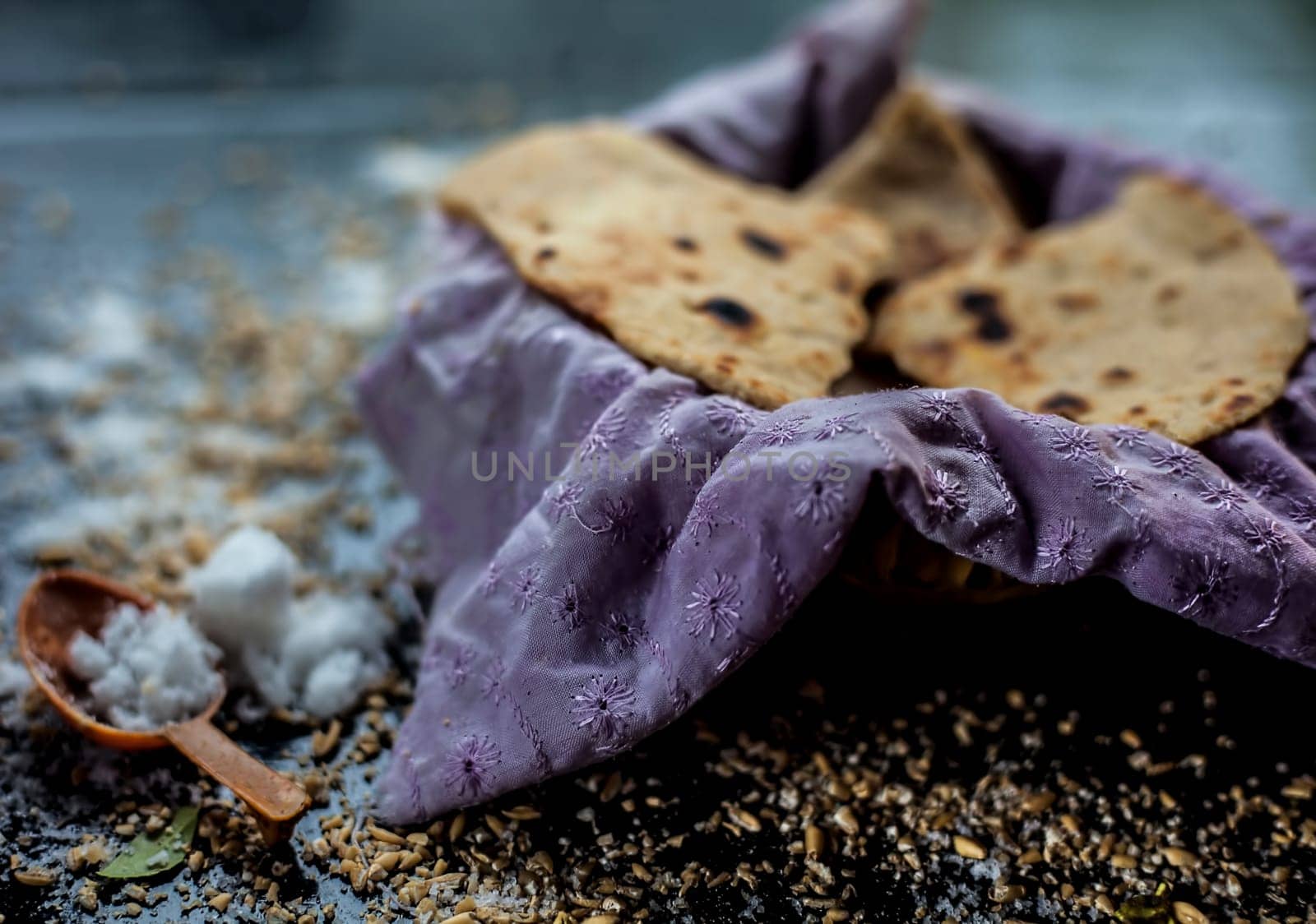 Close-up shot of round bread Bhakri on the black wooden surface along with some raw whole wheat, and salt in a container. Shot of Bhakri in a container on the black surface.