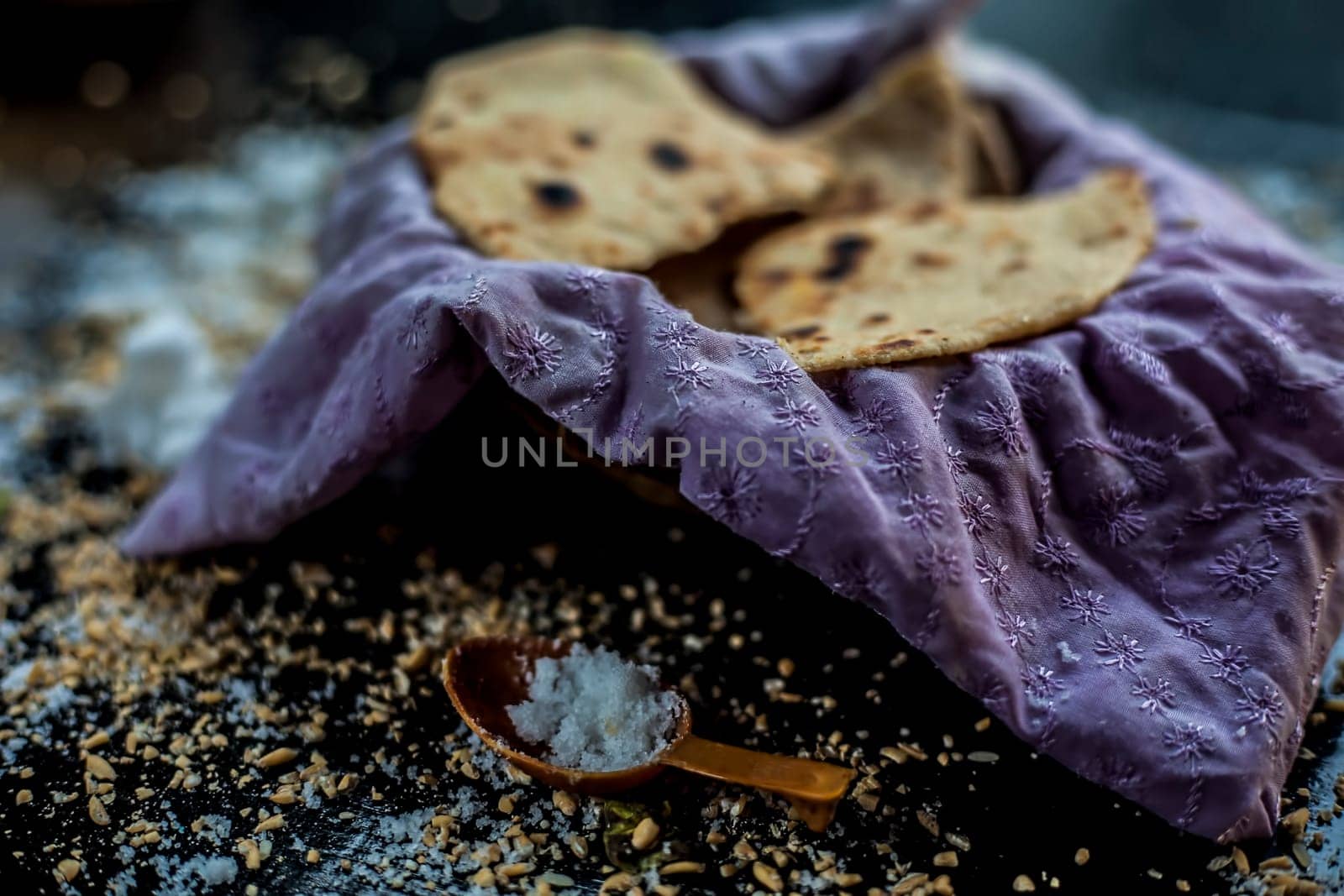 Close-up shot of round bread Bhakri on the black wooden surface along with some raw whole wheat, and salt in a container. Shot of Bhakri in a container on the black surface. by mirzamlk