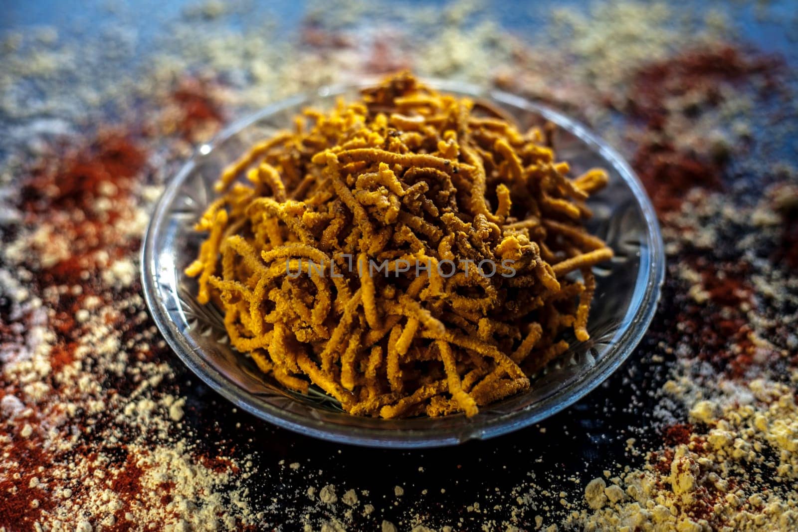 Diwali and Shravan Som special Teekha Gathiya in a glass plate along with some spread chickpea flour, red chili powder, and other ingredients that are needed to make the snack on a black surface.