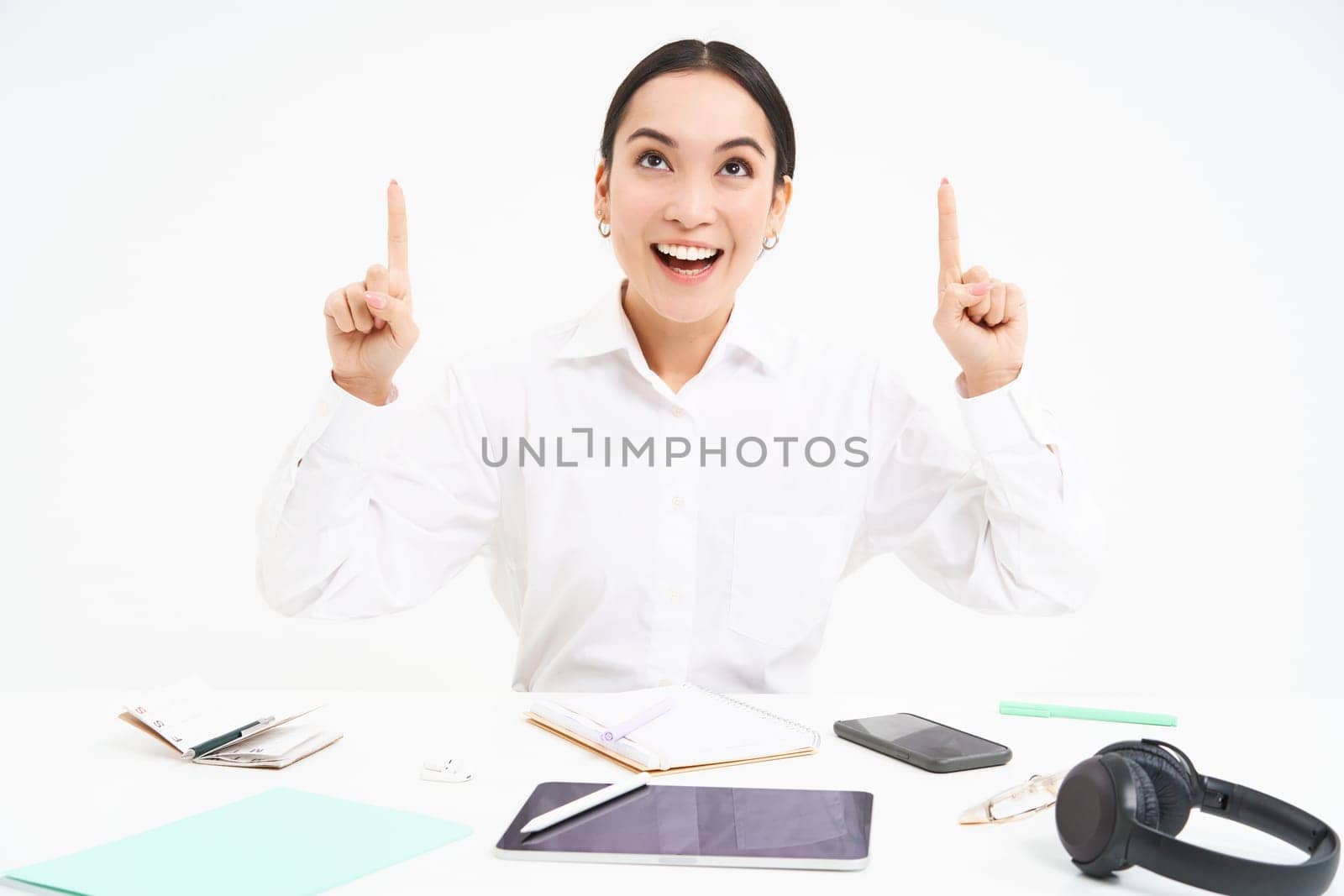 Business and profession. Portrait of asian woman in workplace, sits in office and points up, shows banner advertisement, white background.