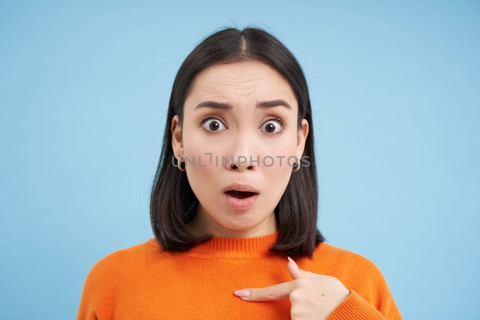Close up of shocked asian girl, points at herself with offended look, stands insulted over blue background by Benzoix