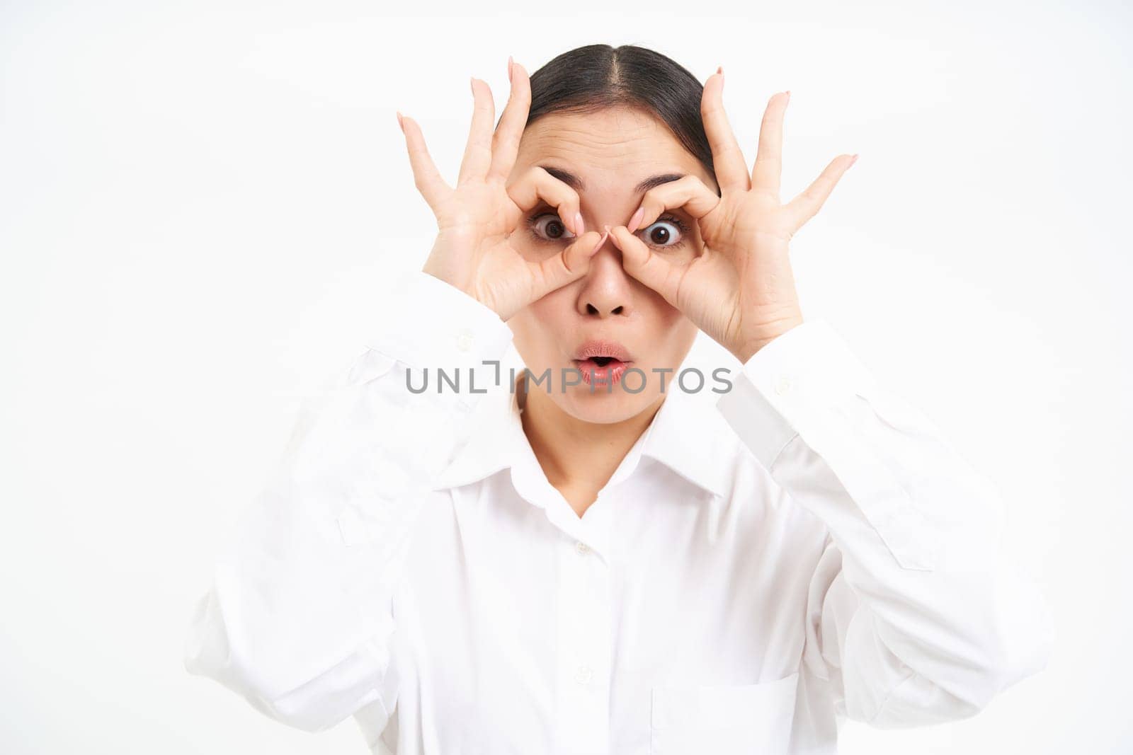 Business people. Happy asian girl looks through finger glasses, stares with surprised face, stands isolated on white background.