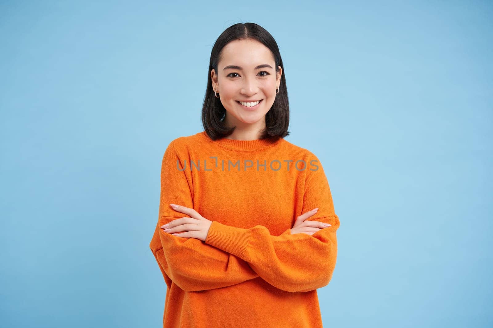 Young asian woman looks with confidence, cross arms on chest and smiles at camera, stands in orange sweatshirt, blue background.