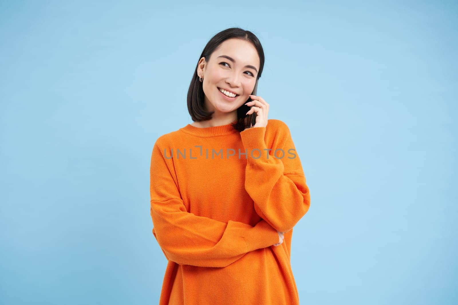 Beautiful smiling asian woman holds mobile phone with happy face. Girl talking on smartphone, listening to voice mail, standing over blue background.