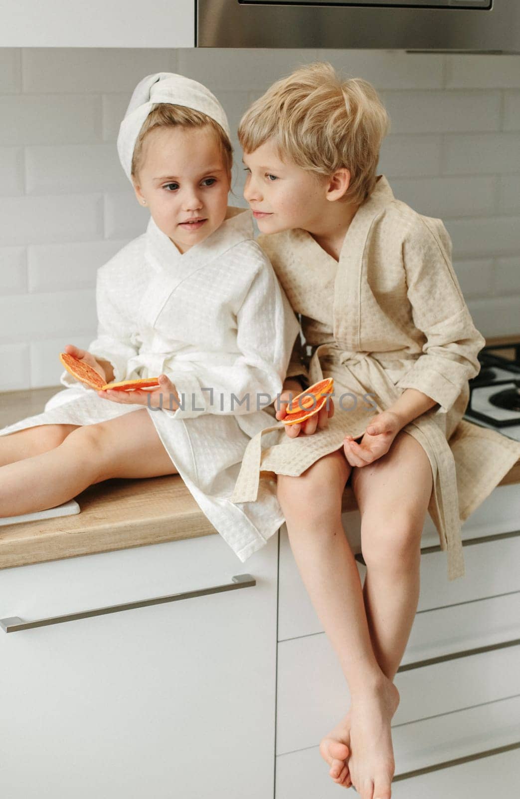 A boy and a girl in bathrobes are sitting in the kitchen talking, eating candied fruit.