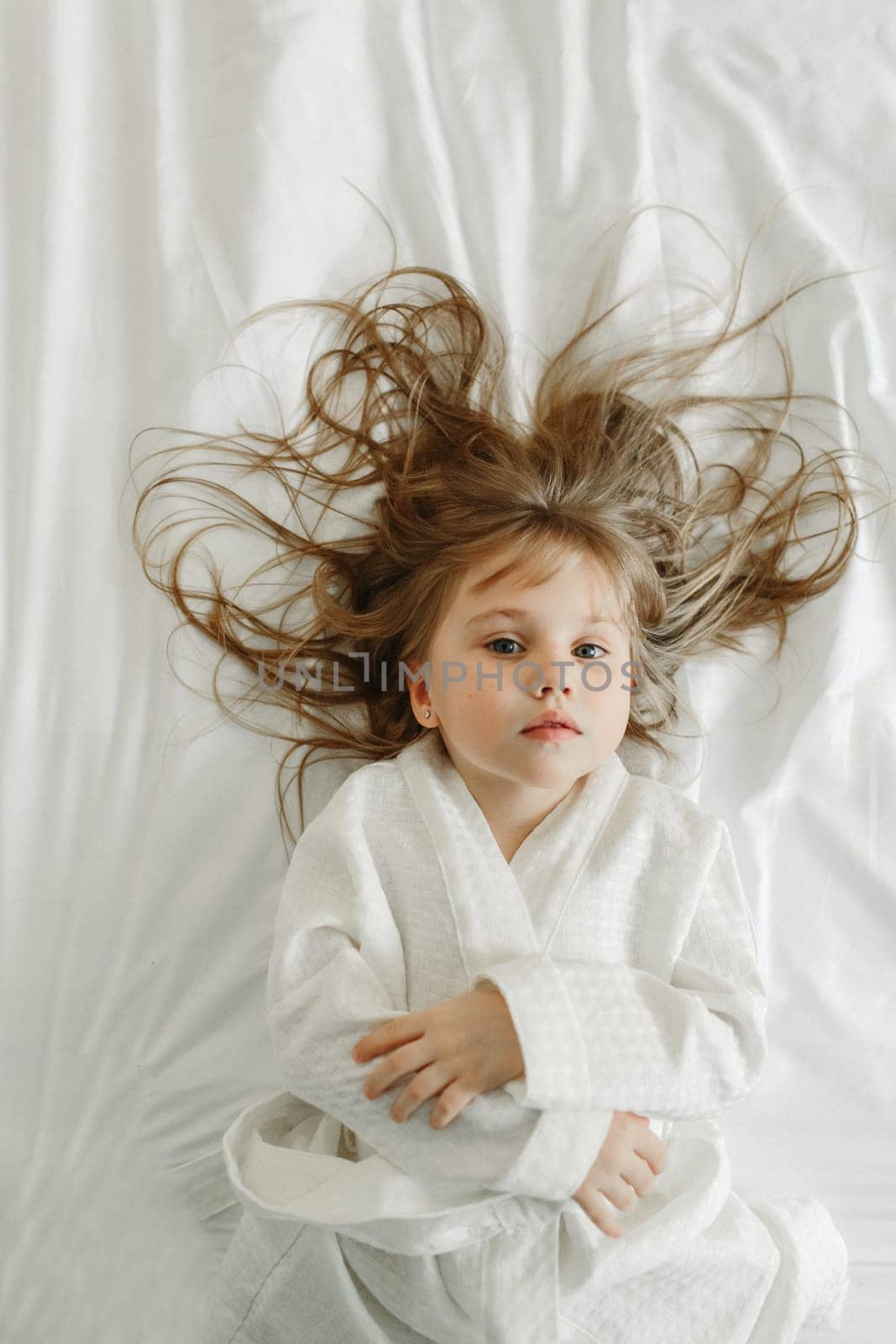 Joyful girl in a white bathrobe lies in bed, looks at the camera. View from above.