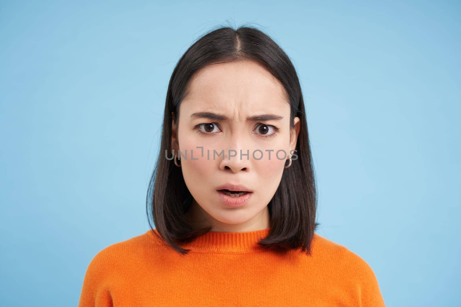 Close up portrait of asian girl with shocked face, looks at smth unbelievable, startled by news, stands over blue background.