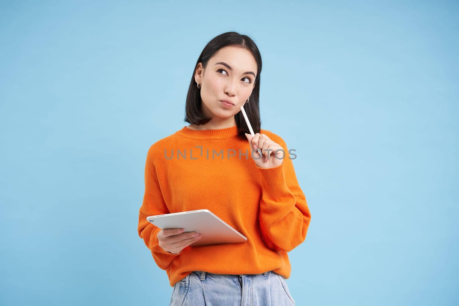 Portrait of young Chinese woman, teacher or student with digital tablet and pencil, writing, taking notes, doing her homework, standing over blue background by Benzoix