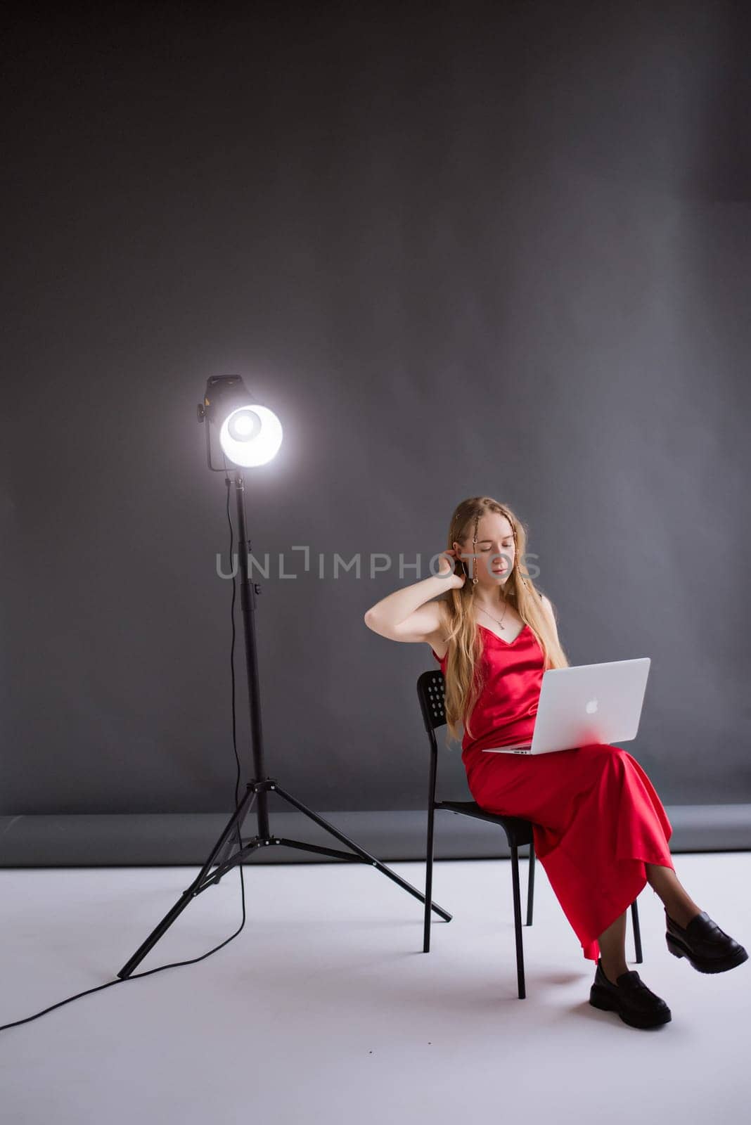 A business woman blogger in a red dress is working, typing on a laptop apple and sitting on a chair, gray background. portrait blonde assistant gaffer of hands with computer MacBook . Vertical