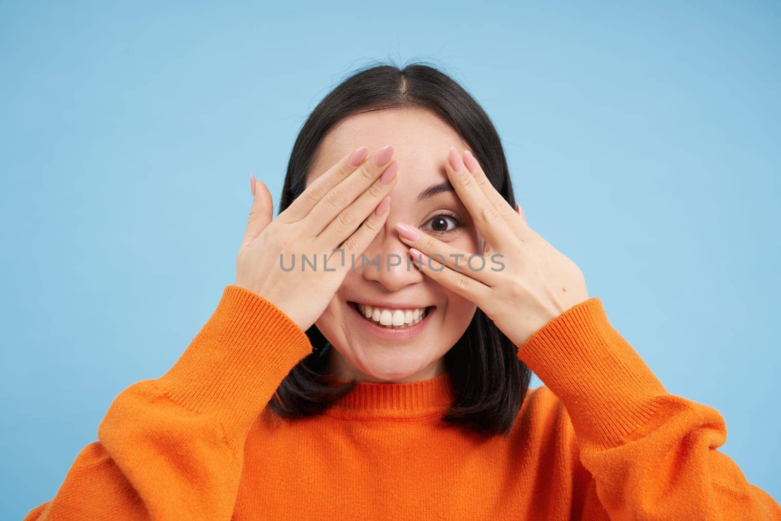 Close up portrait of korean girl waits for surprise, shuts her eyes and peeks through fingers with excited smile, stands over blue background.