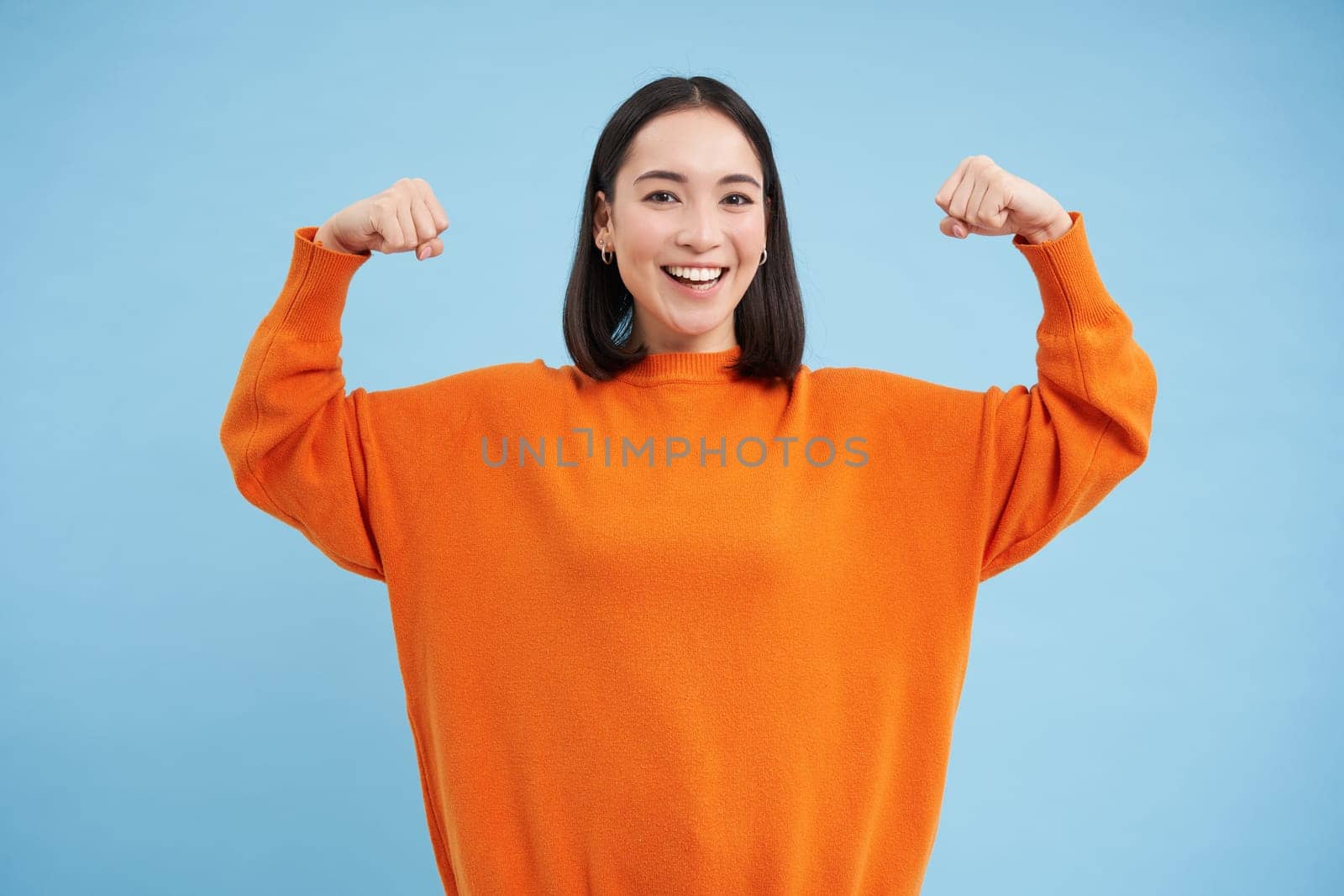 Strong and healthy people. Young asian woman shows her muscles, flexing biceps and looks confident, smiling pleased, in great shape, blue background by Benzoix