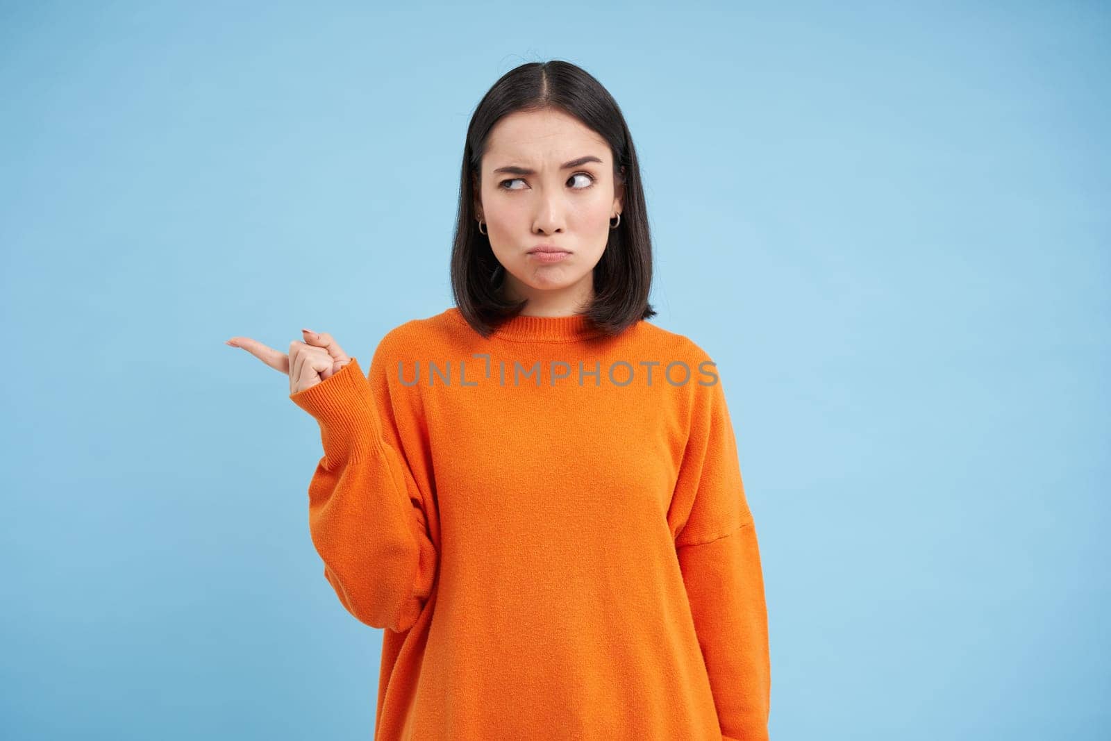 Confused korean girl cant understand something, points left with clueless face, thinking, standing in orange shirt against blue background.