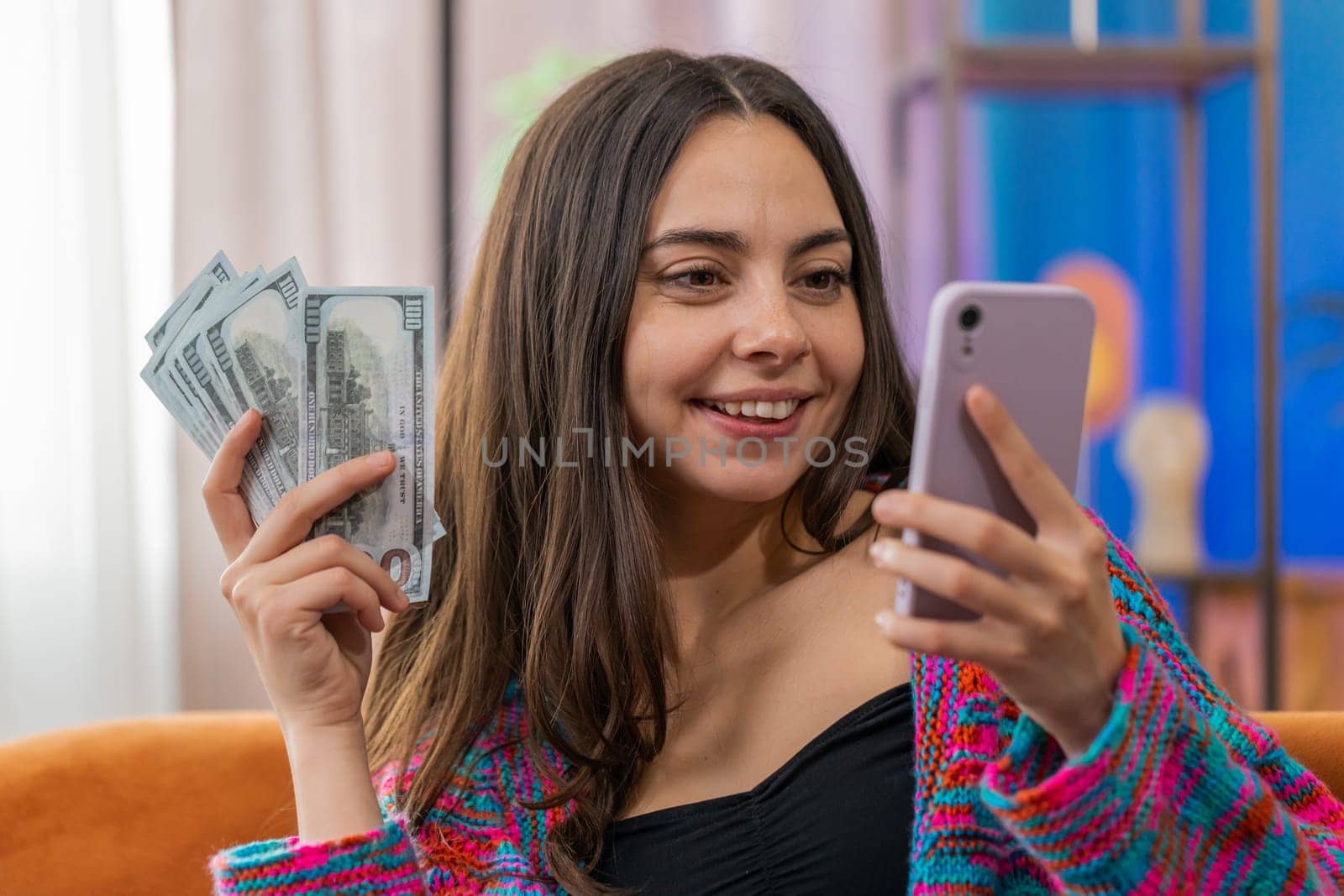 Planning family budget. Woman counting money cash, use smartphone calculate domestic bills. Joyful girl satisfied of income and saves money for planned vacation, gifts at home apartment room on sofa