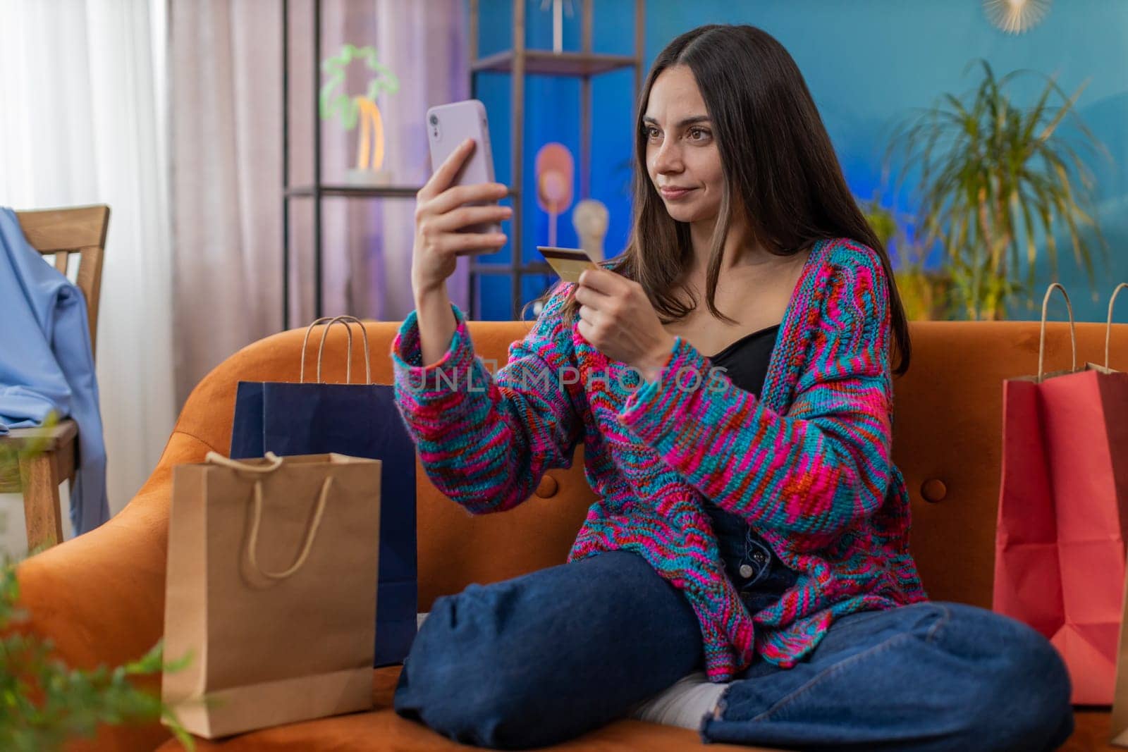 Happy shopaholic consumer young woman sitting with shopping bags making online order, payment with credit plastic bank card. Discount sale on internet. Portrait of girl at home apartment room on sofa