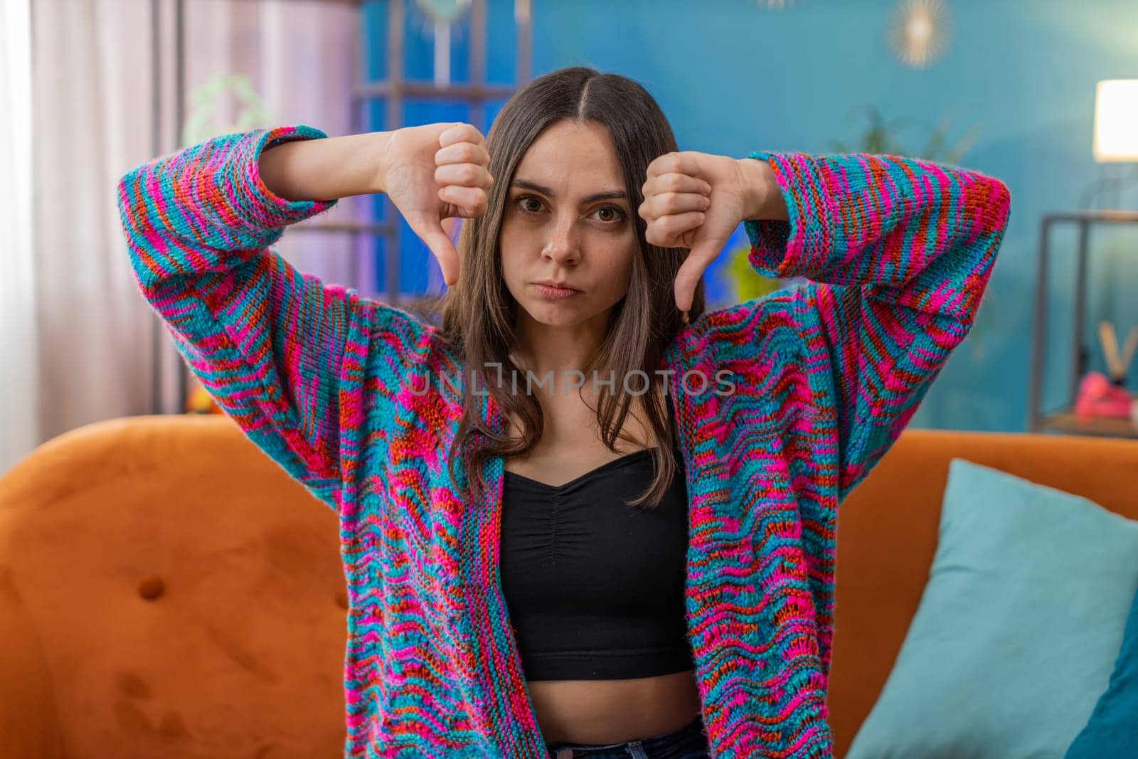 Dislike. Upset young woman showing thumbs down sign gesture, expressing discontent, disapproval dissatisfied bad work at modern home apartment indoors. Displeased Caucasian girl in living room on sofa