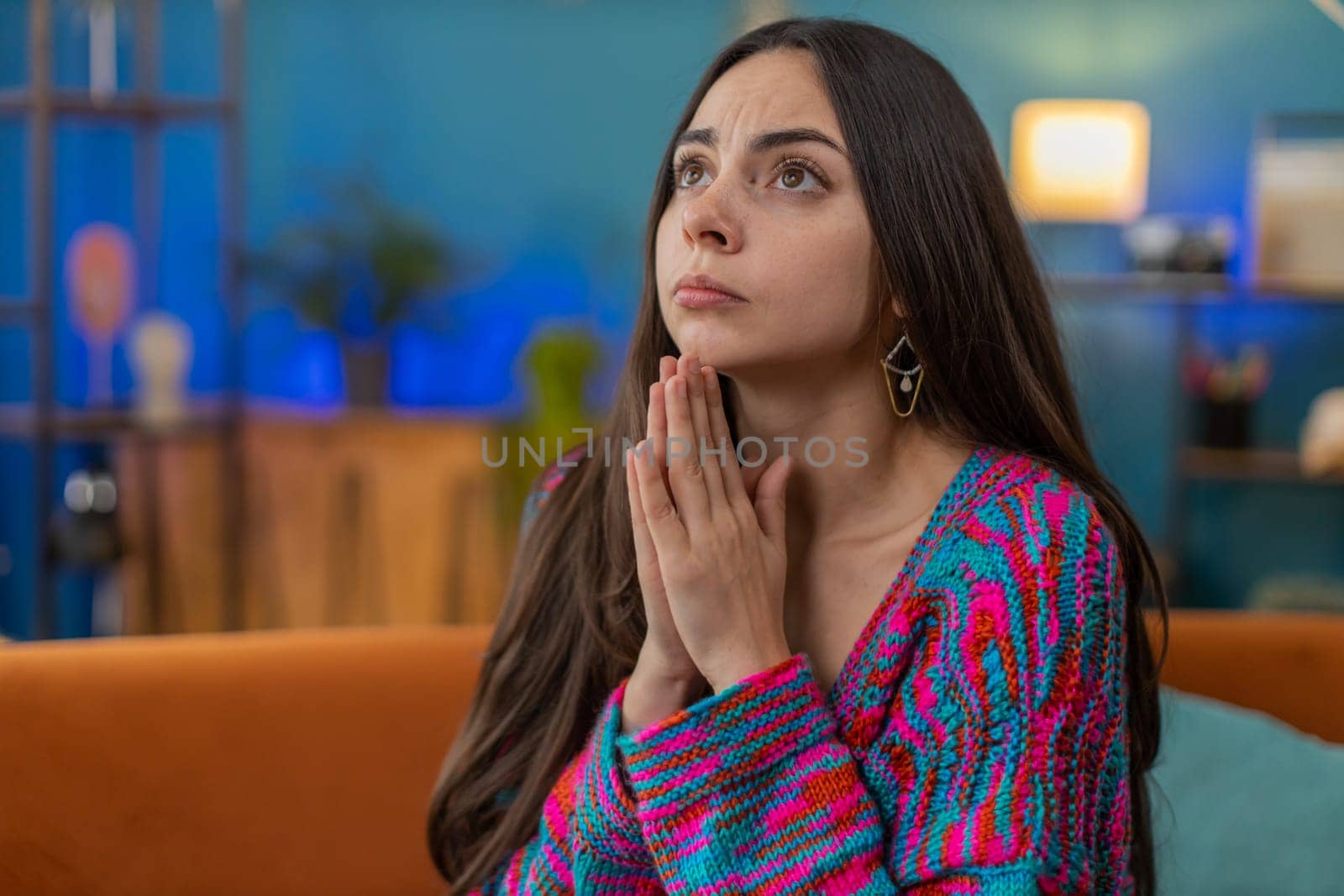 Please, God help. Young woman praying sincerely with folded arms, looking upward and making wish, asking with hopeful imploring expression, begging apology. Sad girl at home apartment room on sofa