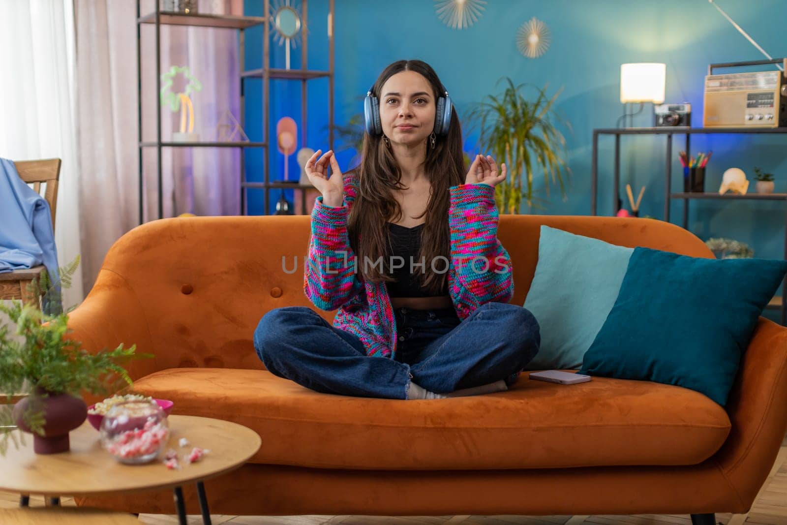 Keep calm down, relax, inner balance. Young woman breathes deeply with mudra gesture, eyes closed meditating with concentrated thoughts, peaceful mind. Portrait of girl at home apartment room on sofa