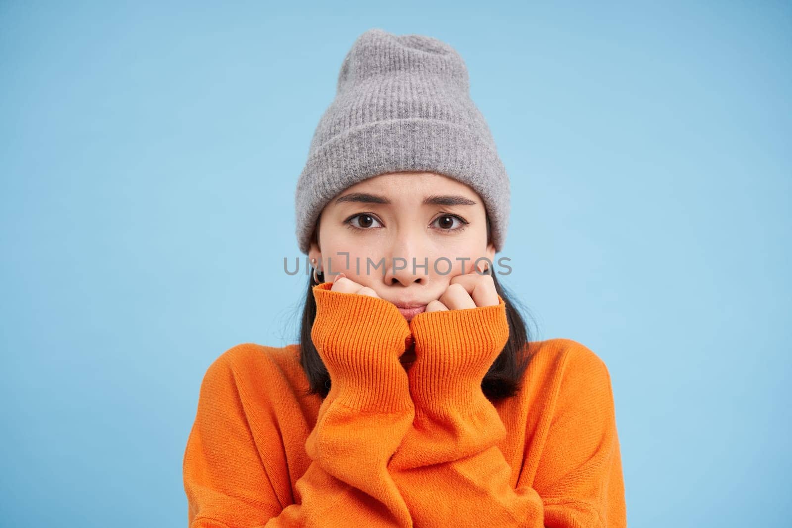 Portrait of asian girl in warm hat, looks with fear, shaking and trembling scared, standing frightened against blue background.