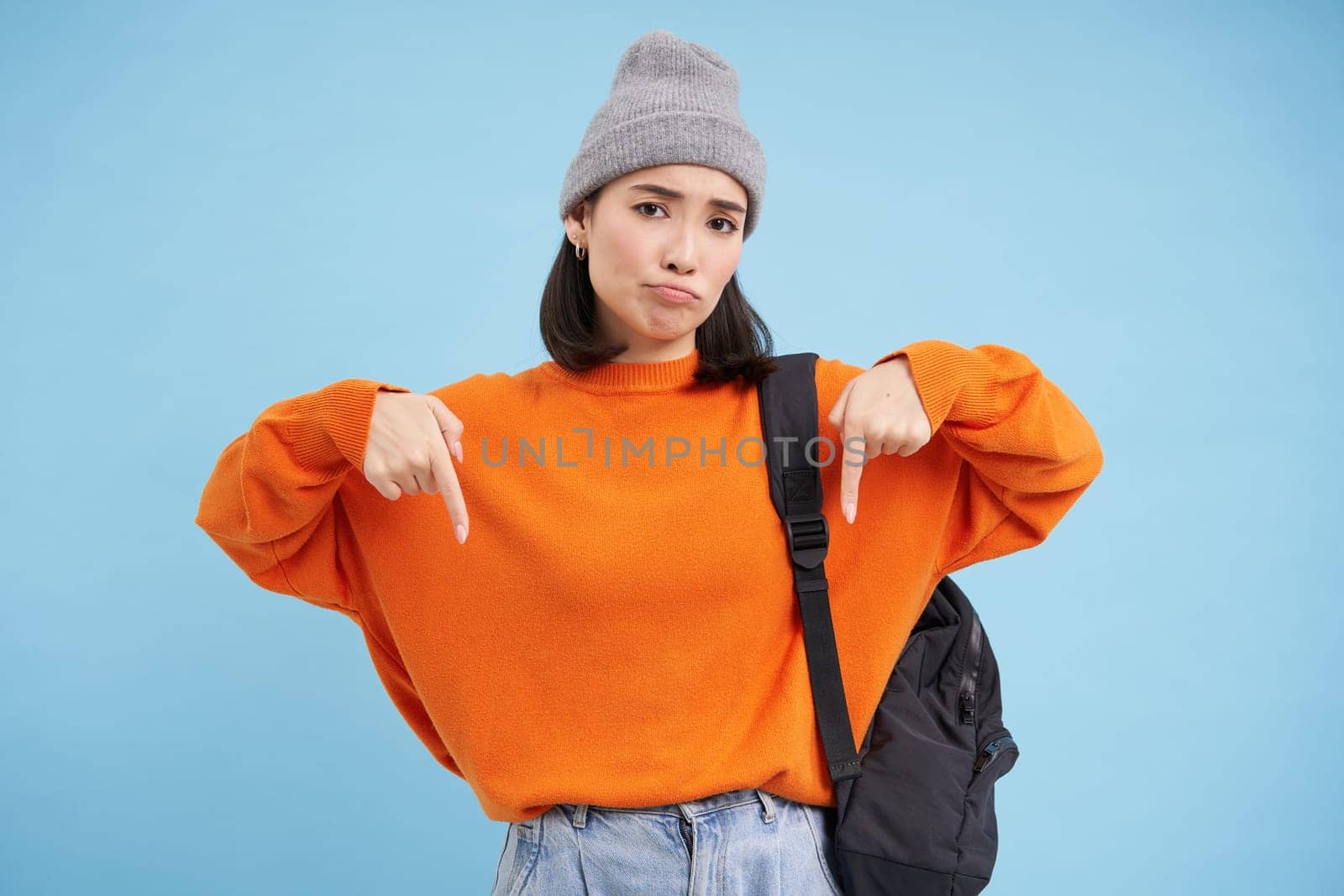 Asian woman in warm hat, wears street outfit and backpack, points fingers down, shows advertisement below, standing over blue background.