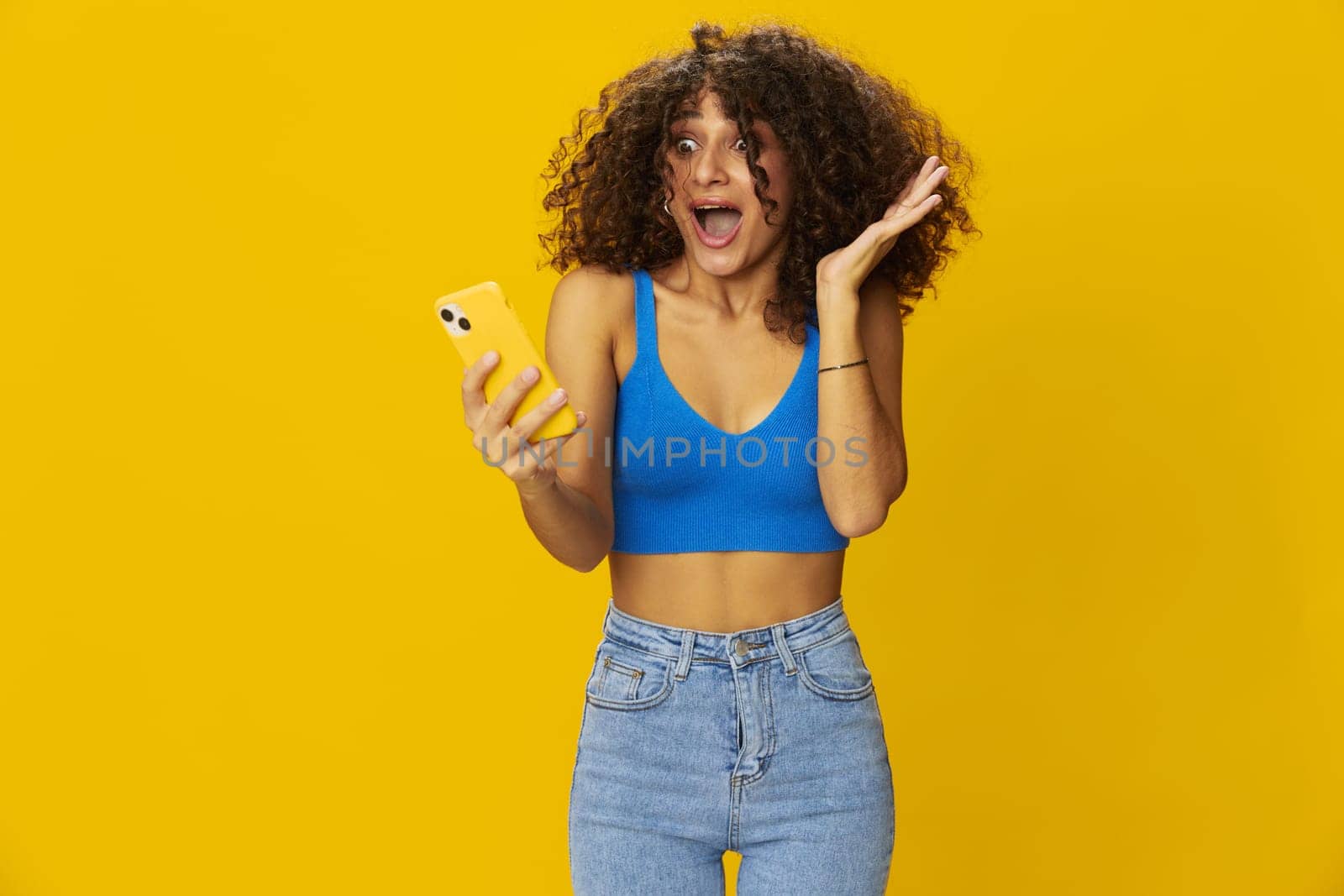 Influencer woman with phone in hand taking selfies talking on video call freelancer social media technology communication at work, with curly afro hair in blue t-shirt on yellow background , smile with teeth and happiness, copy space. High quality photo