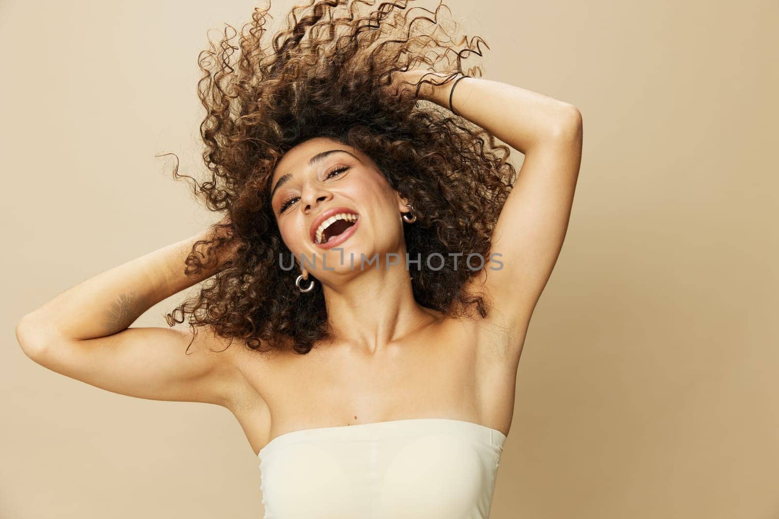 Woman applies cream and balm to her curly hair, the concept of protection and care, a healthy look, a smile with teeth on a beige background.