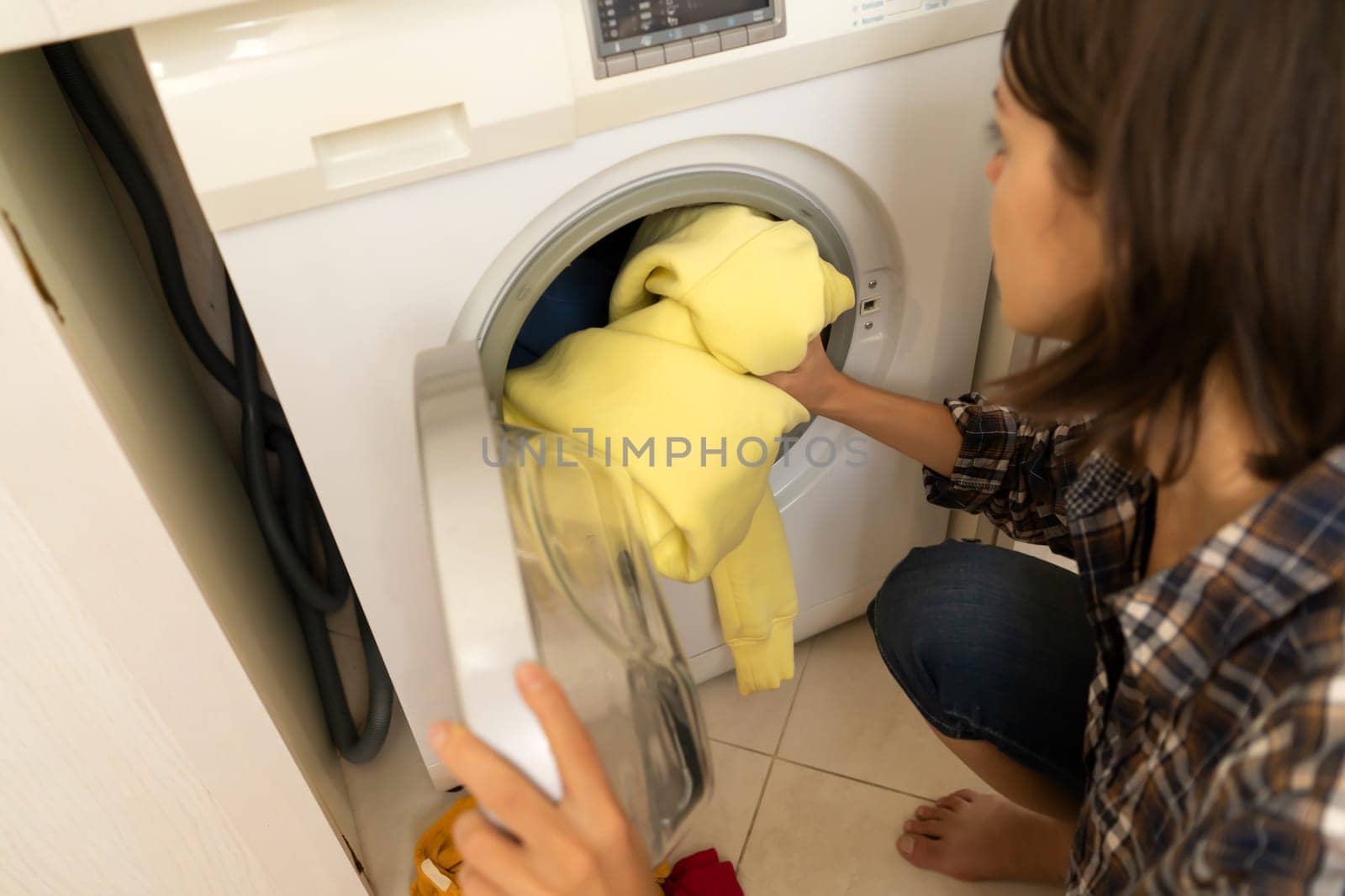 A girl folds bright clothes into a new washing machine in the bathroom at home, a woman washes things and spin them out with detergent in the laundry close-up, top view.