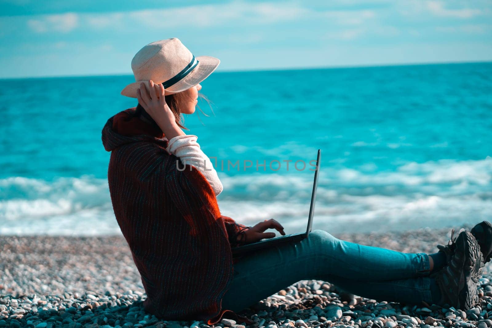 Young girl in a light hat and casual clothes sits on the beach by the sea with a laptop on a sunny day, works, studies, buys tickets during a trip, a woman rests on vacation and types on the keyboard.