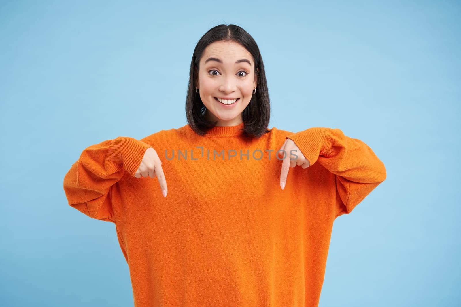 Excited asian woman pointing down, shows advertisement below with happy smile, look there gesture, stands over blue background by Benzoix