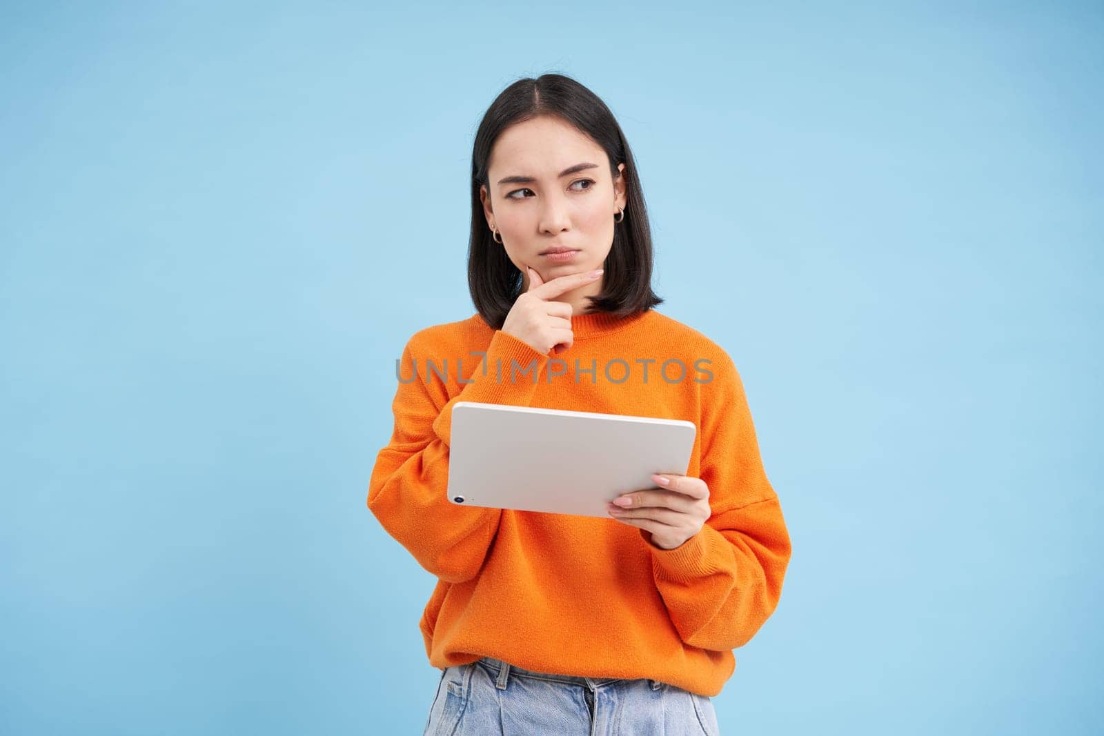 Woman with digital tablet thinking, looking aside with thoughtful face expression, standing over blue background by Benzoix