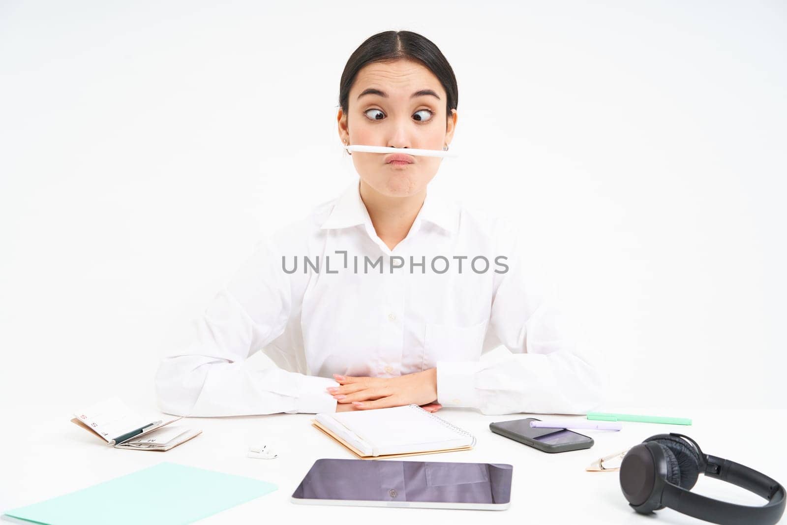 Business and office work. Young woman sitting at workplace, fooling around, holds pen with lips and shows funny faces, white background.