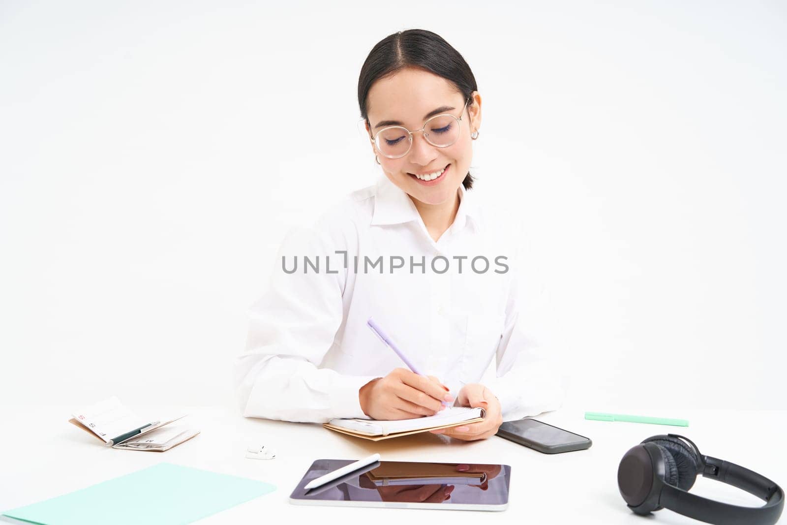 Young asian office woman, sitting at workplace desk, making notes, smiling and looking professional, white background.