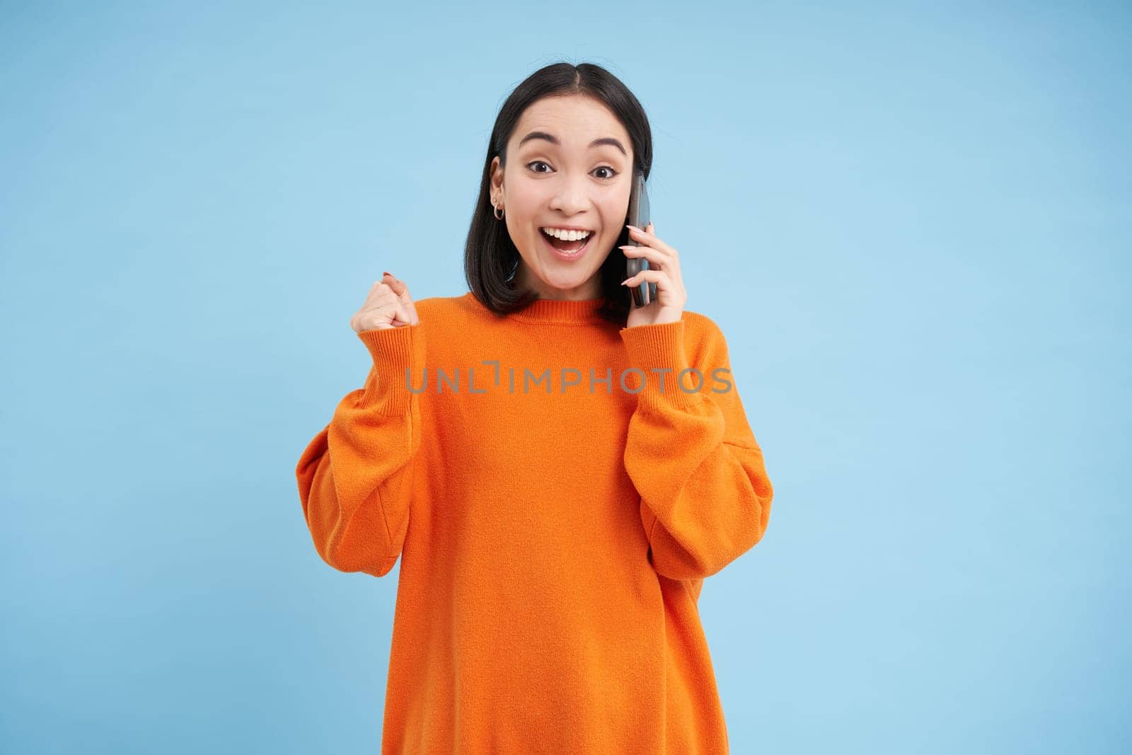 Surprised korean woman answers phone call and reacts to great news, celebrating, standing over blue studio background by Benzoix