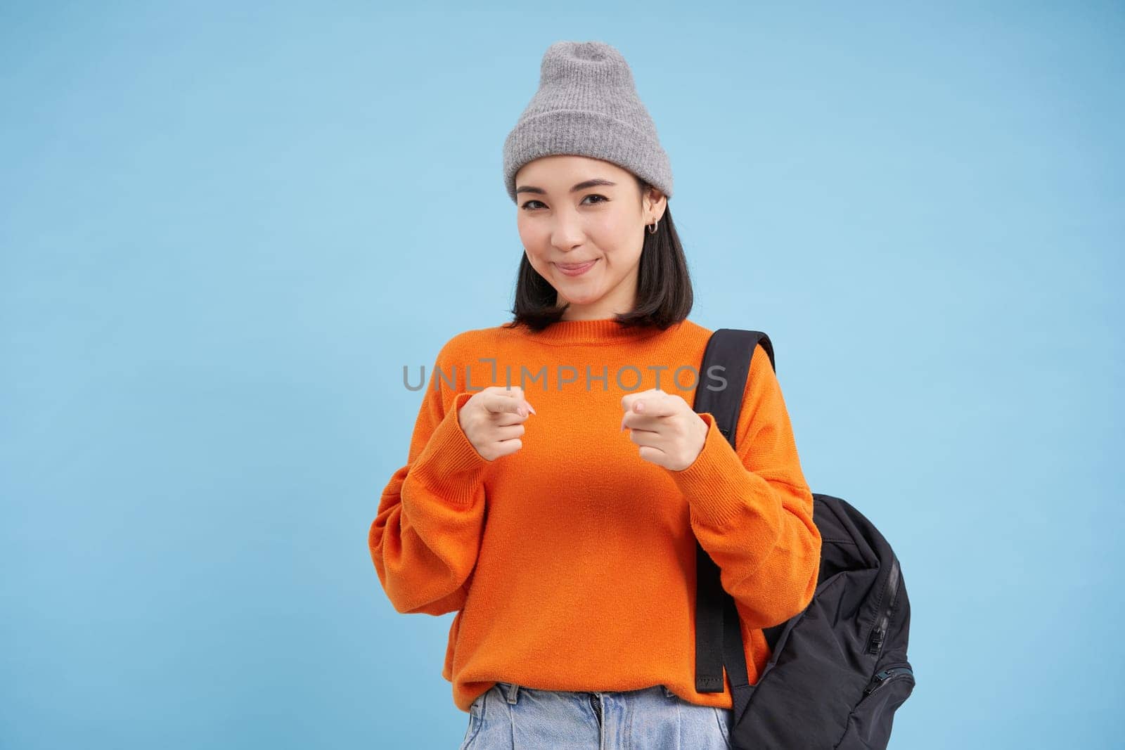 Cute korean woman in hat with backpack, student pointing fingers at camera, inviting you, congratulating, standing over blue background.