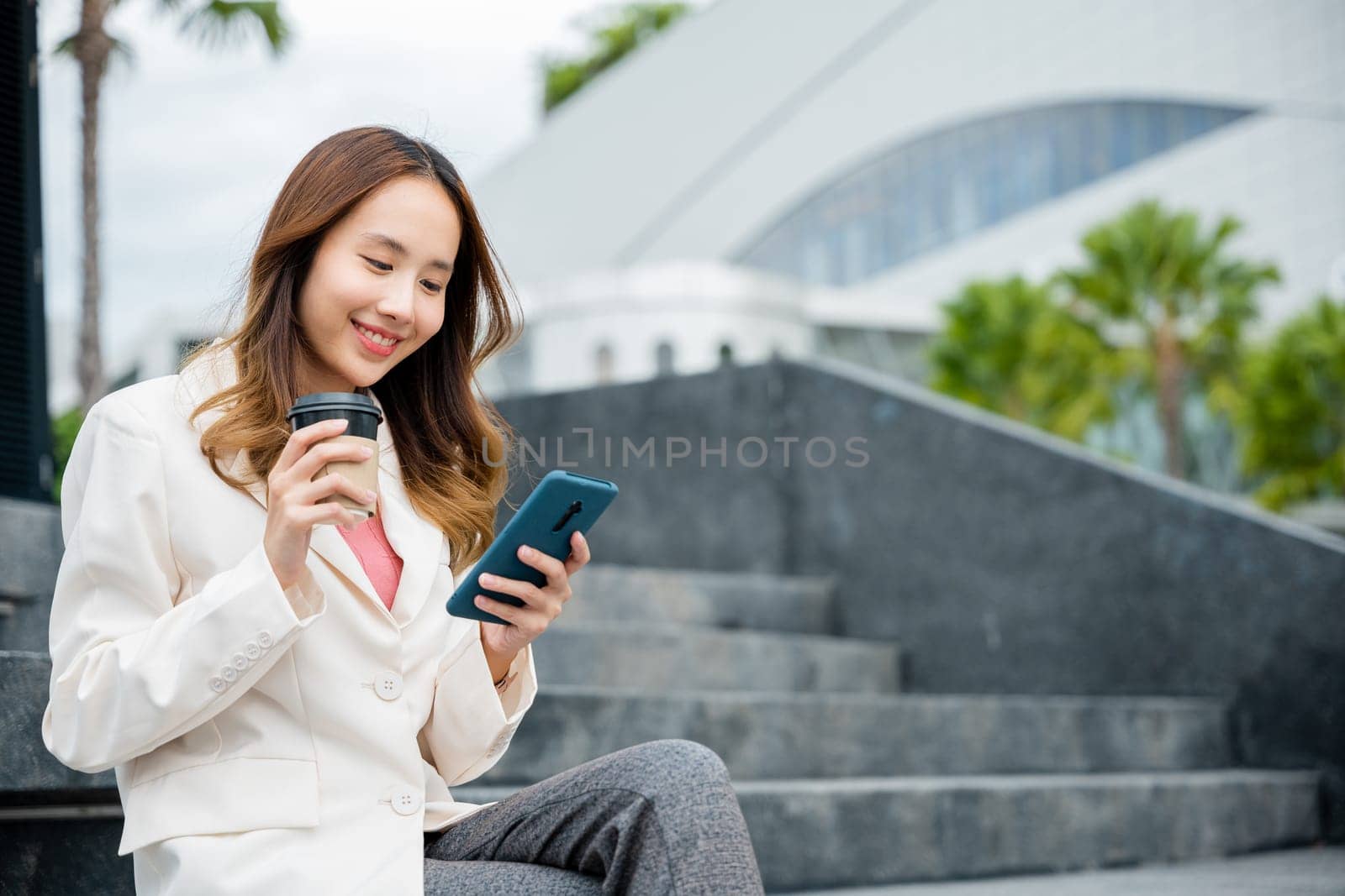 Young Asian woman wearing blue shirt while use cellphone with holding coffee cup sitting on staircase. Female business people using smart phones in the city outdoors.