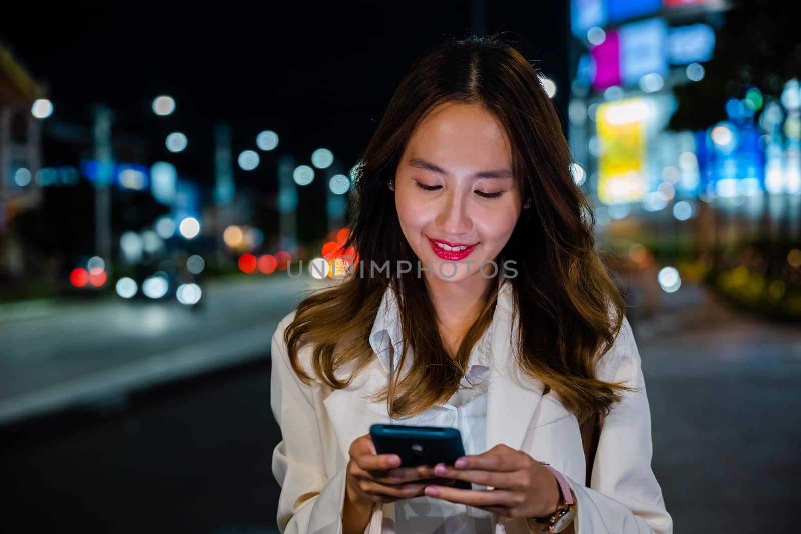 Business woman using mobile phone walking through night city street while waiting car to pick up home, Beautiful young smiling female texting work on smartphone, social media