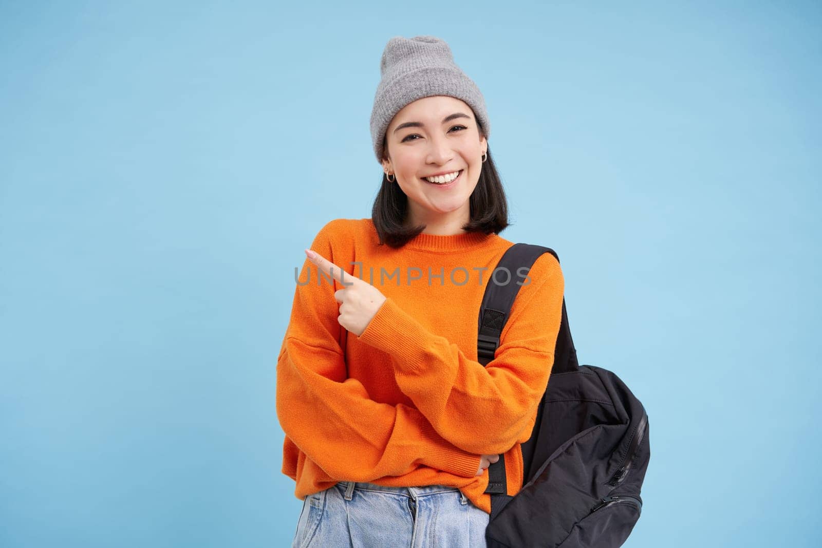 Stylish korean woman in hat, holds backpack, points finger at advertisement, shows promo offer banner, stands over blue background.