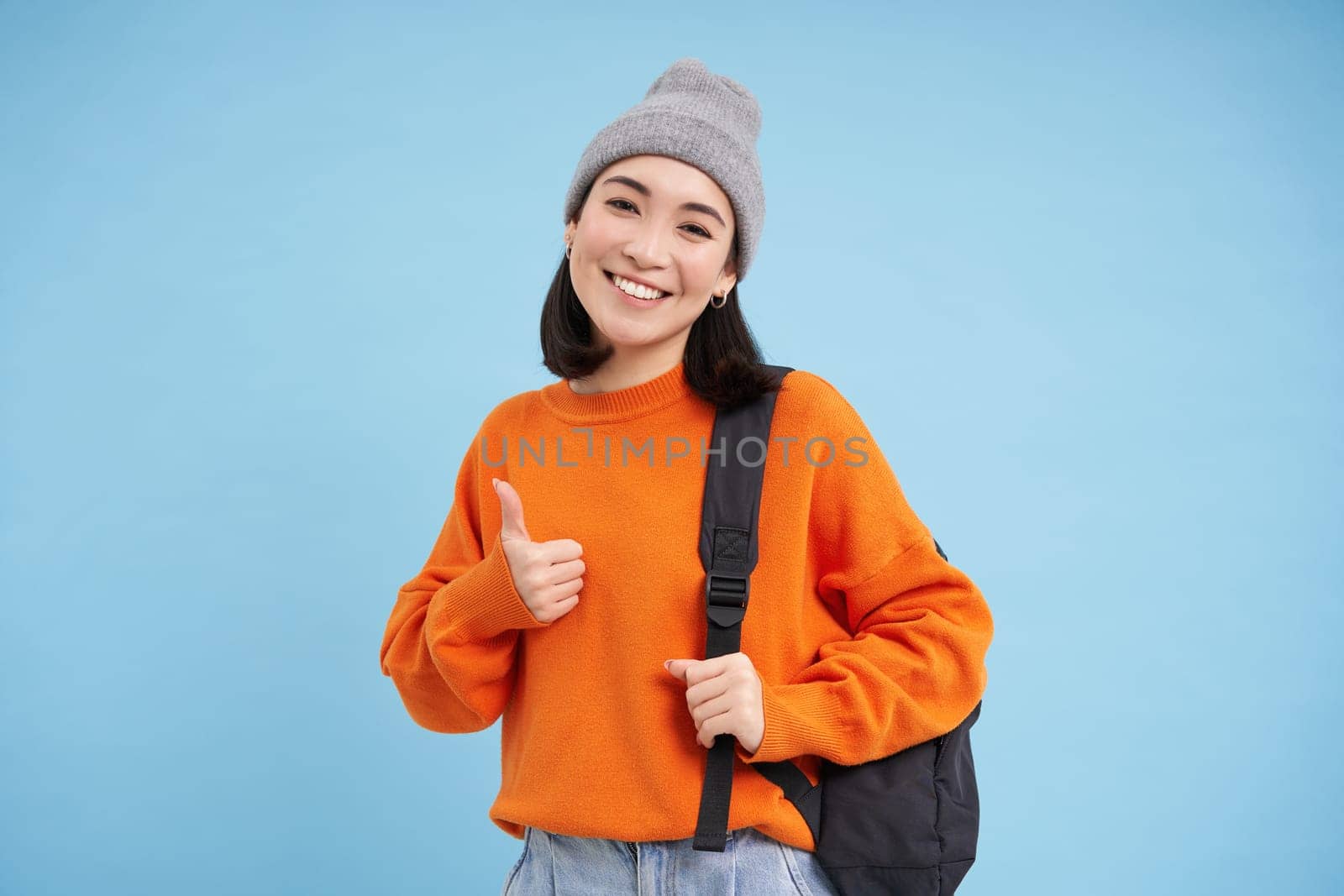 Happy young student, asian woman in warm hat, holds backpack, shows thumbs up and smiles, blue background.