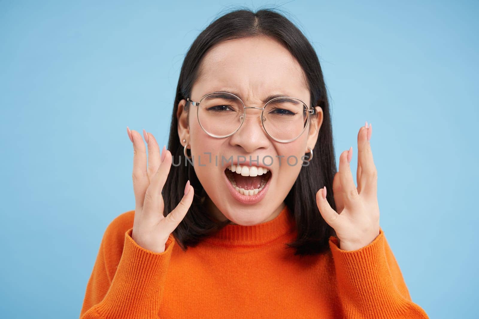 Portrait of angry japanese girl in glasses, screaming and looking furious, shaking hands in anger, looks outraged, stands over blue background by Benzoix