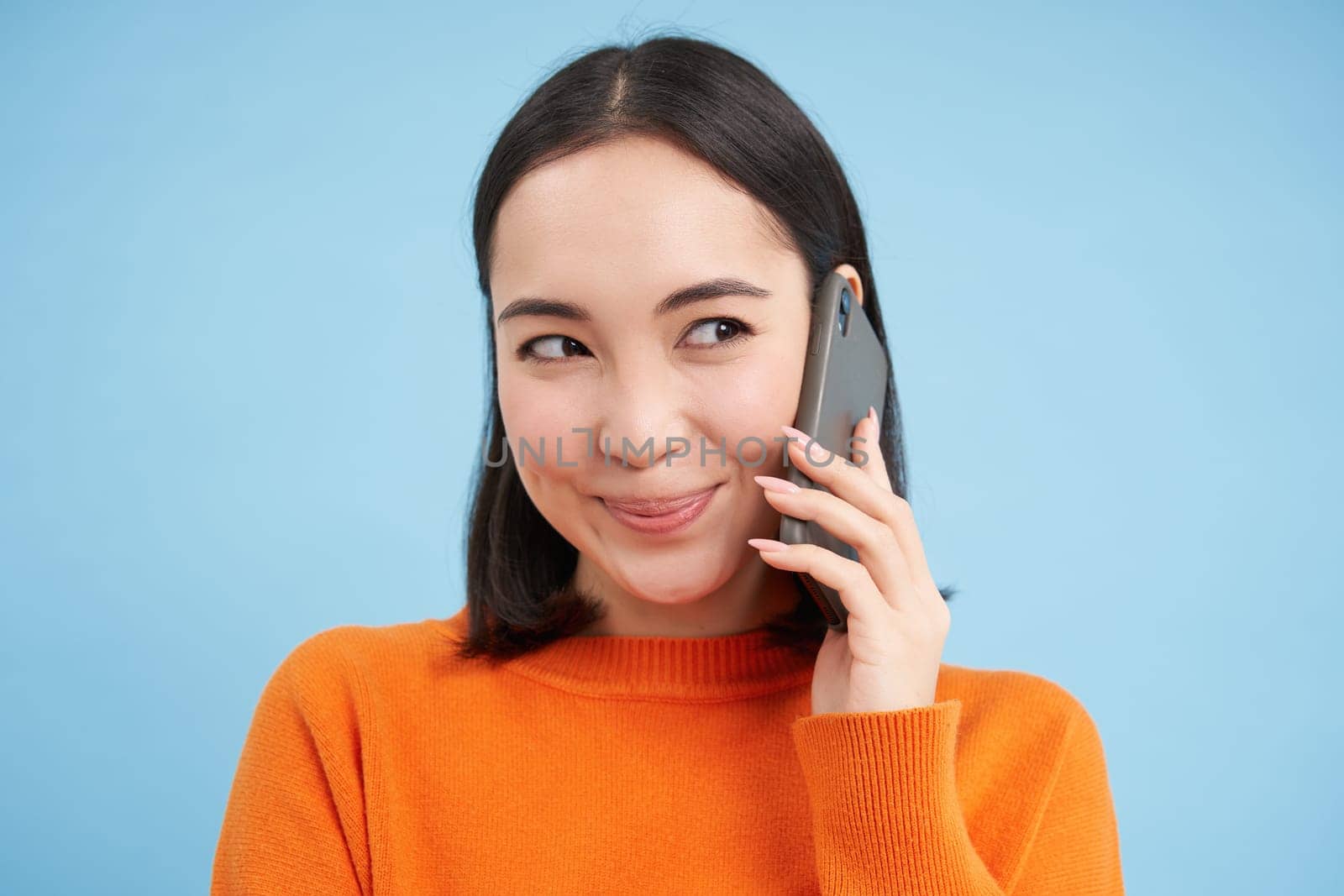 Technology concept. Close up of smiling asian woman talks on mobile phone, having conversation on cellphone, blue background by Benzoix