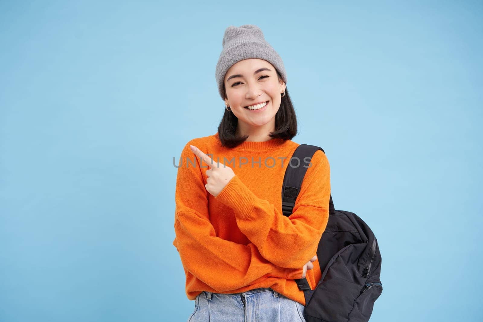 Beautiful smiling korean woman in hat, pointing finger left, showing information banner, demonstrates offer, stands over blue background by Benzoix