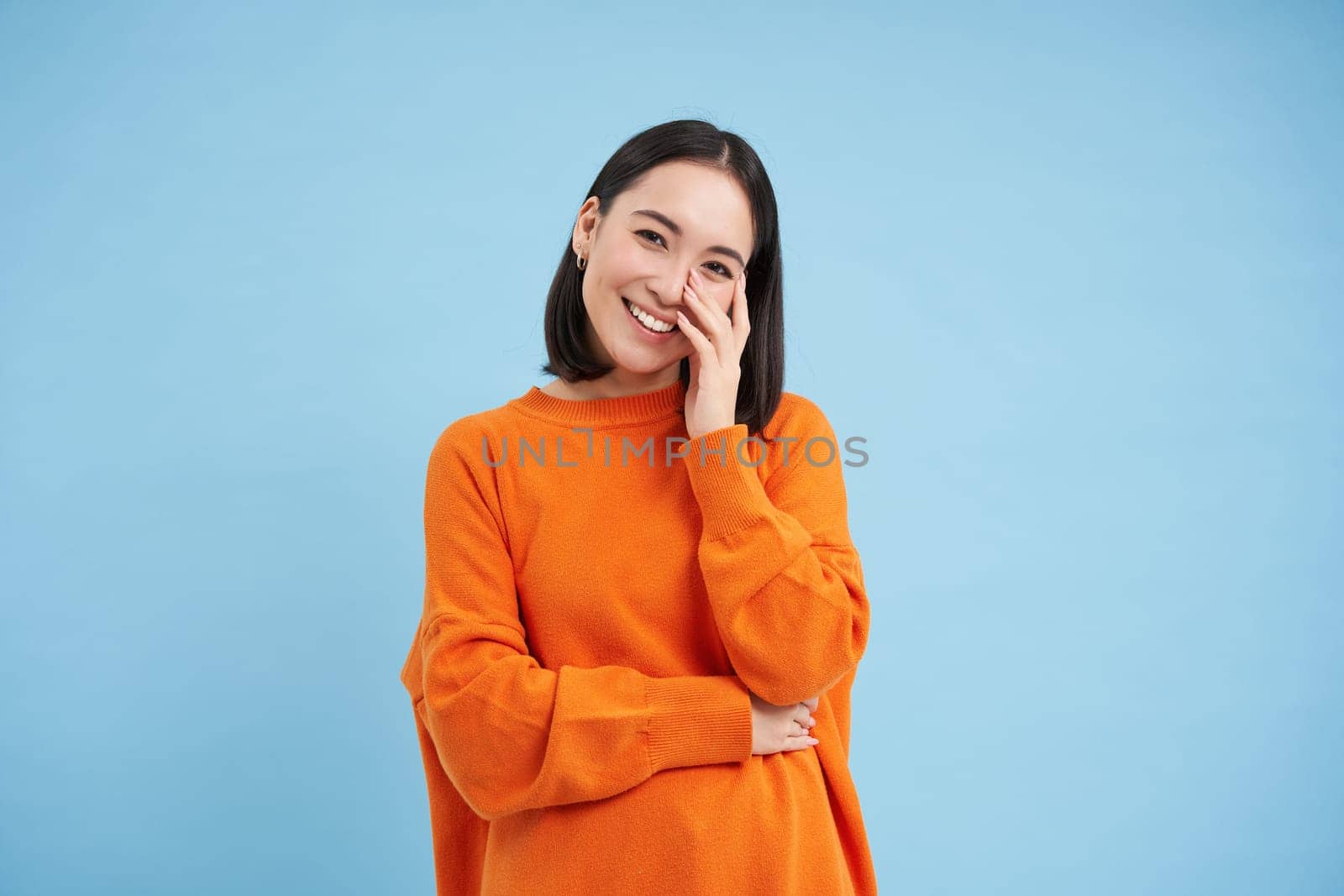 Portrait of young happy woman in orange jamper, touches her clear glowing skin, has beautiful white smile and candid emotions, blue background.