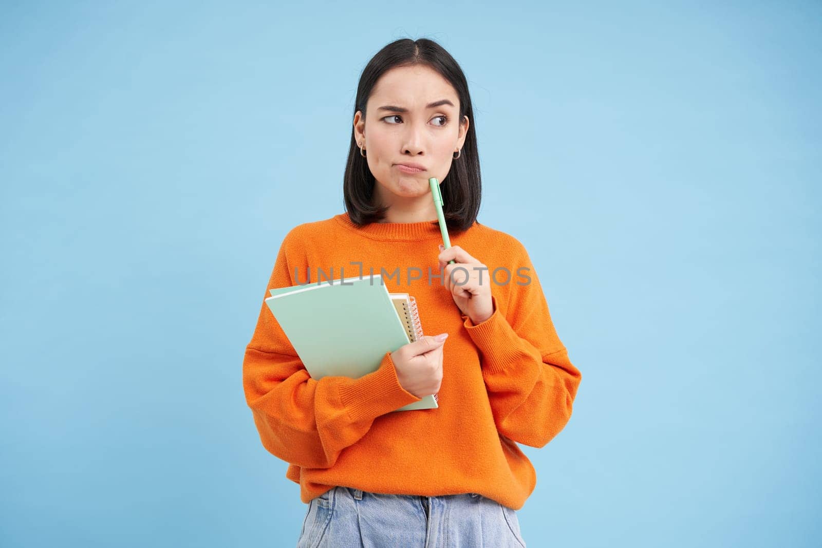Asian girl, student with thinking face, pondering smth with serious face, holding pencil and notebooks, standing over blue background by Benzoix