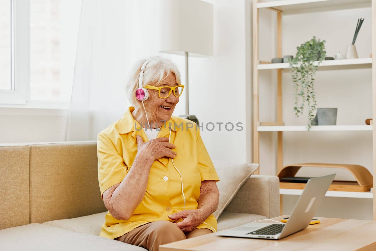 Elderly woman wearing headphones with a laptop communication online video call smile happiness, sitting on the couch at home and working in a yellow shirt, the lifestyle of a retired woman. High quality photo